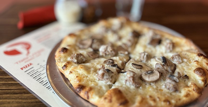 A mushroom and cheese pizza on a metal tray with a restaurant menu underneath, on a wooden table.