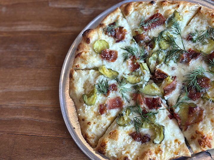 A pickle and bacon pizza topped with fresh dill, served on a metal tray on a wooden table.