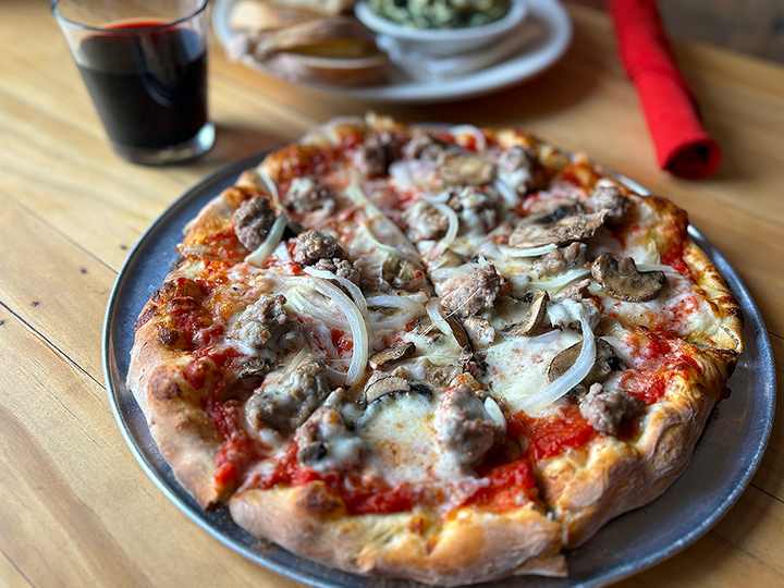 A pizza with sausage and onion slices on a metal tray, with a glass of red wine and bowls of salad in the background on a wooden table.