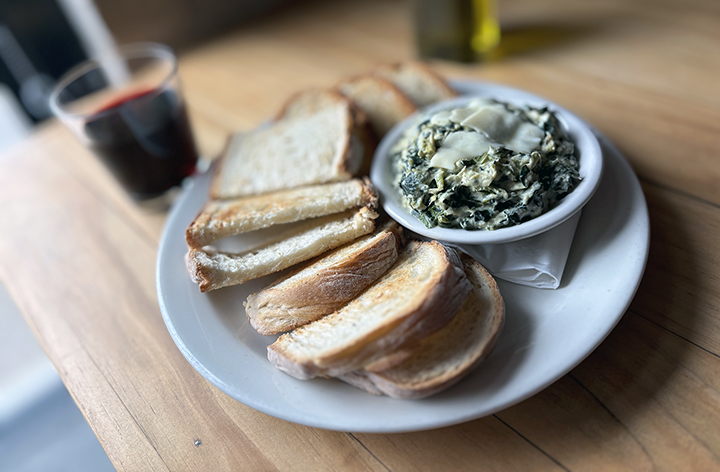 Plate of toasted bread slices with spinach and cheese dip, glass of red wine, and a glass of water on a wooden table.