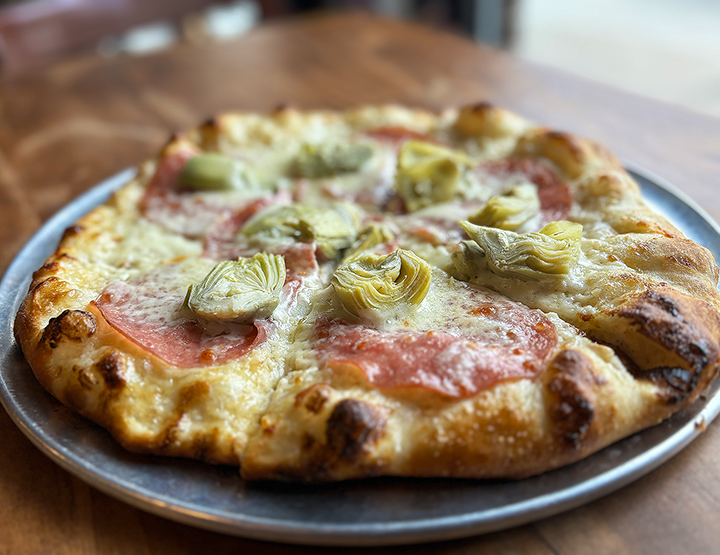 Close-up of a pizza with cheese, ham, and artichoke toppings on a metal plate placed on a wooden table.