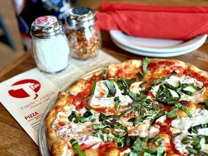 Close-up of a pizza with tomato sauce, cheese, and basil on a metal tray at a wooden table, with salt, crushed red pepper, and red napkins in the background.