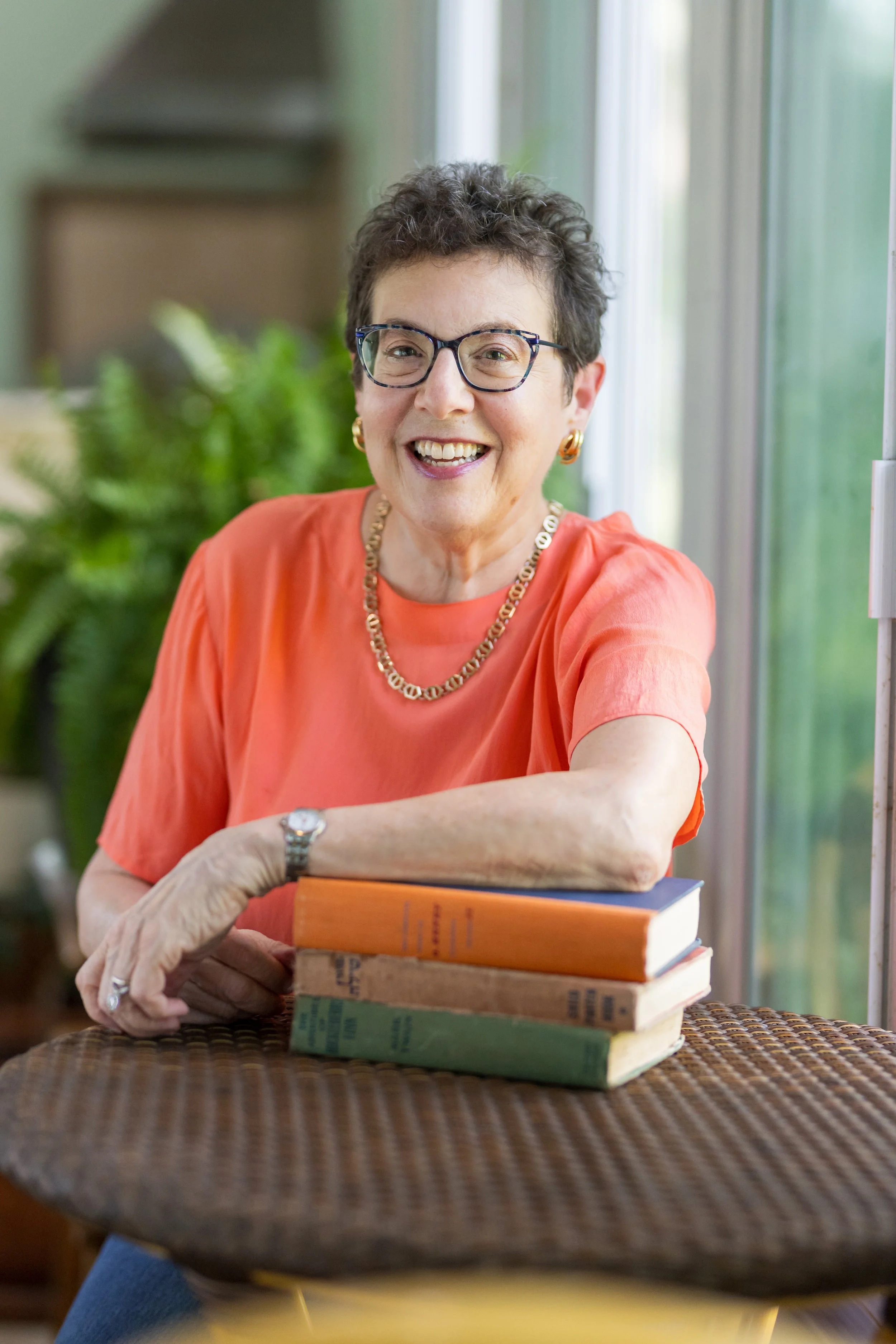 Smiling elderly woman with short curly hair, glasses, and gold jewelry, sitting at a table with stacked books, wearing a coral-colored top.