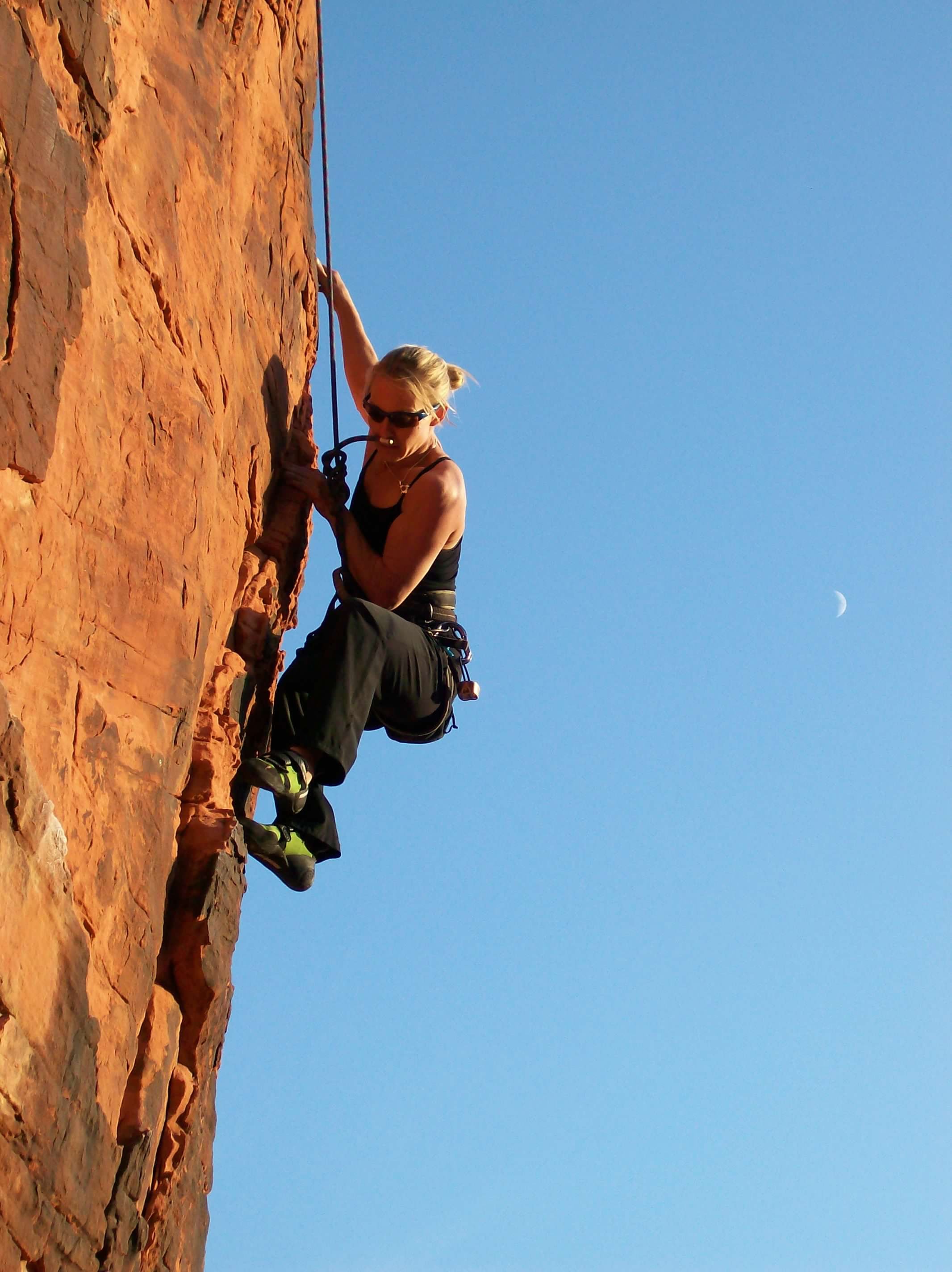 Dr. Sandy rock climbing on a steep red rock face, wearing climbing gear and sunglasses, against a bright blue sky with visible moon.
