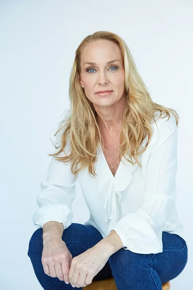 Dr. Sandy with long blonde hair, fair skin, and blue eyes, wearing a white blouse and blue jeans, sitting against a plain white background.
