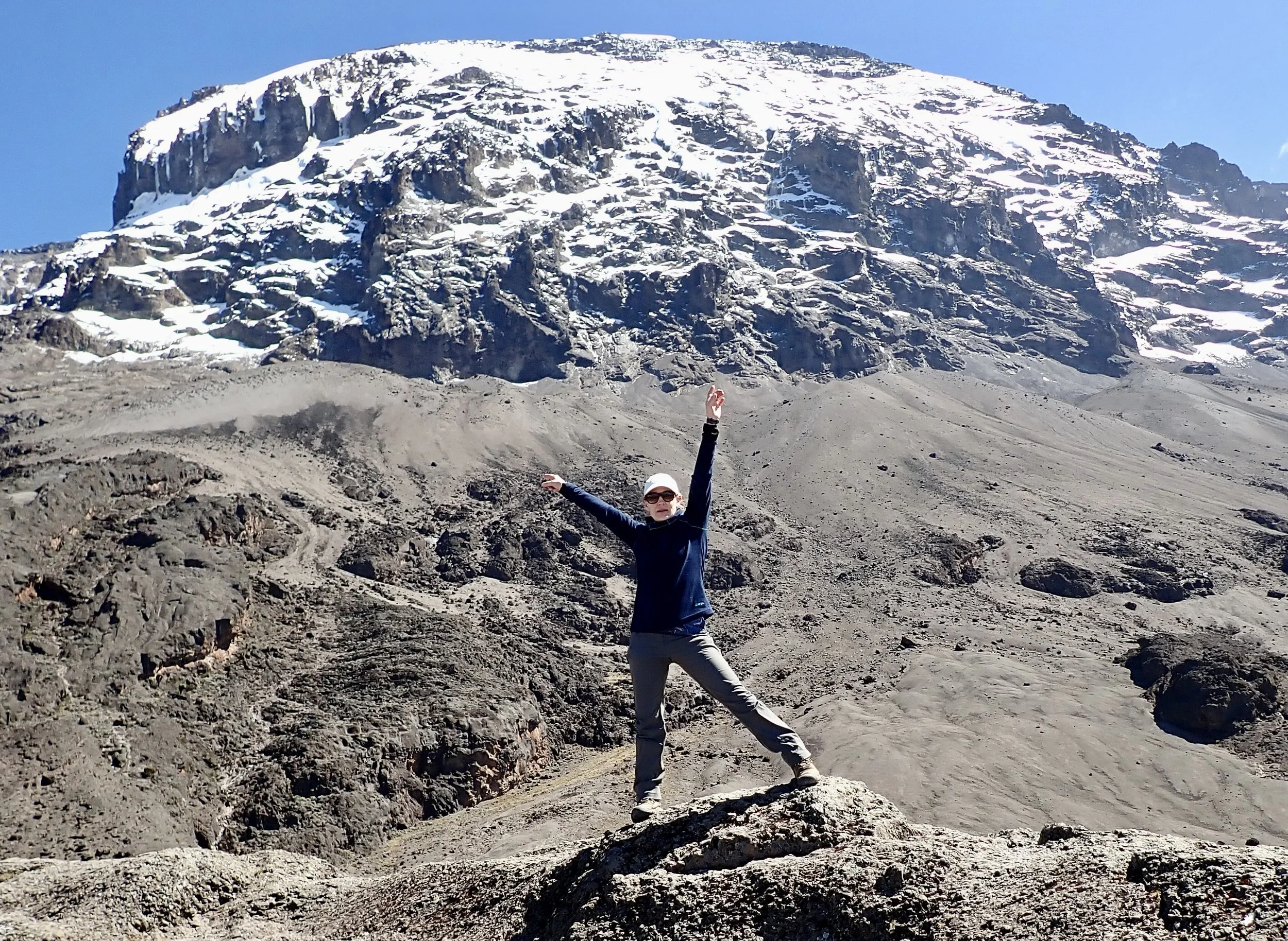 Dr. Sandy standing on rocky terrain with arms raised, mountain with snow and glacier in background.