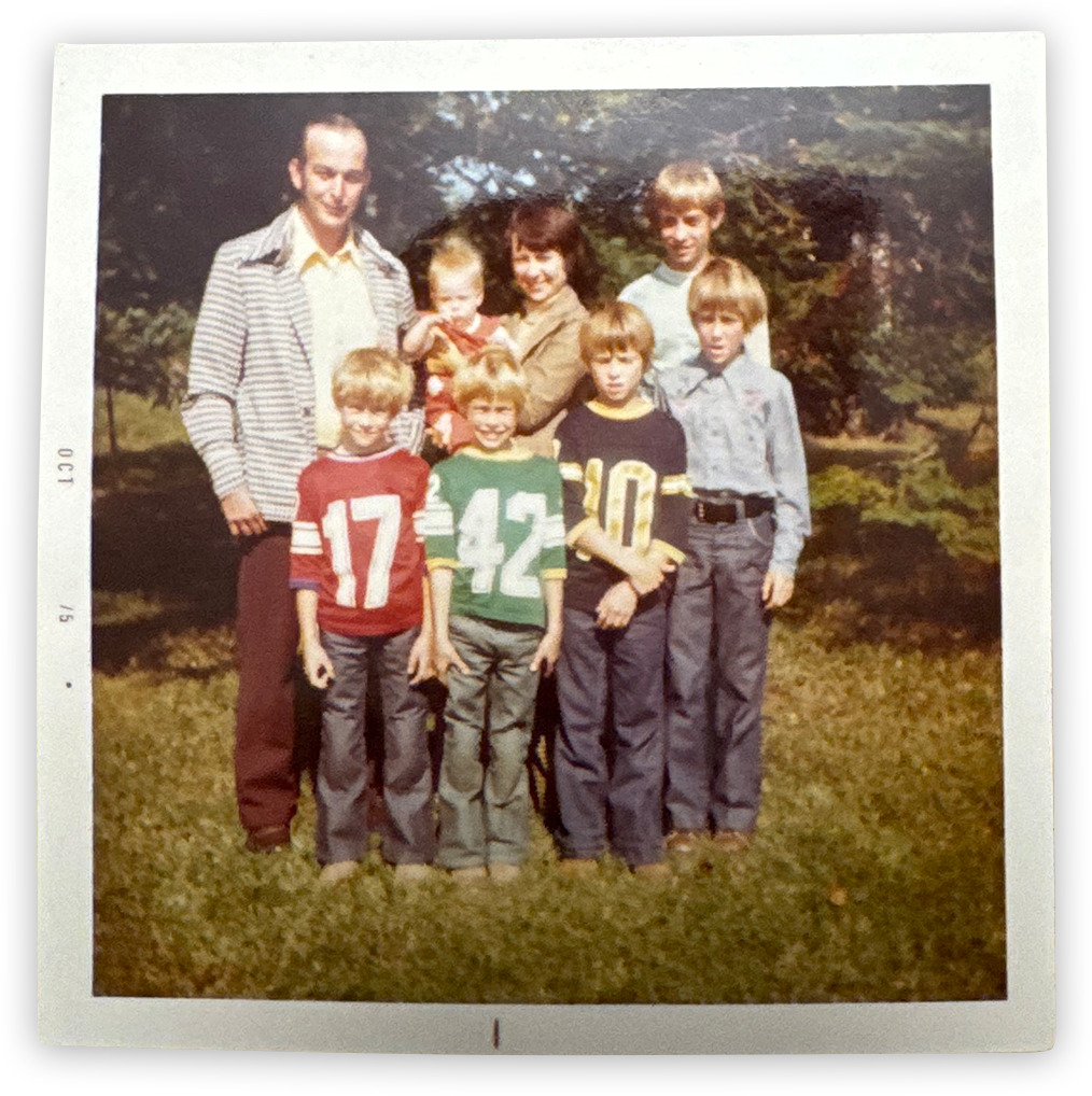 Family portrait of two adults and six children outdoors on grass, some wearing football jerseys, with trees in the background.