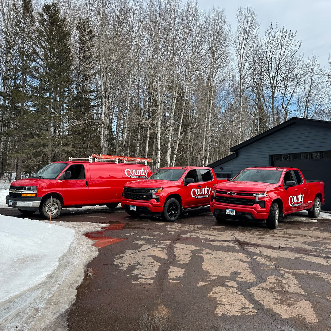 Three red County Pluming utility vehicles parked on a driveway in front of a black garage, with snow and leafless trees in the background.