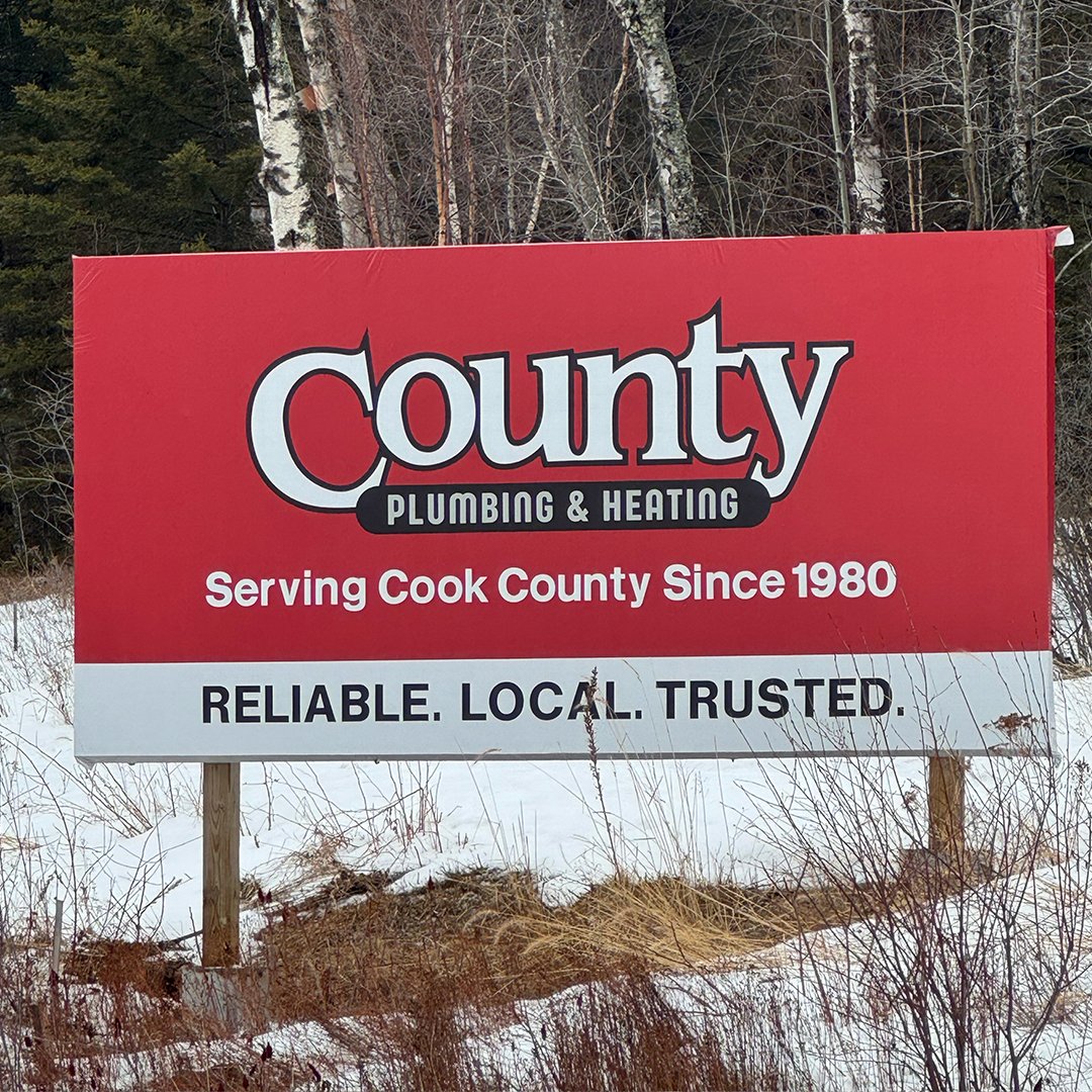 A large red and white outdoor sign on two wooden posts reads 'County Plumbing & Heating' and 'Serving Cook County Since 1980' with the slogan 'Reliable. Local. Trusted.' in black text below.