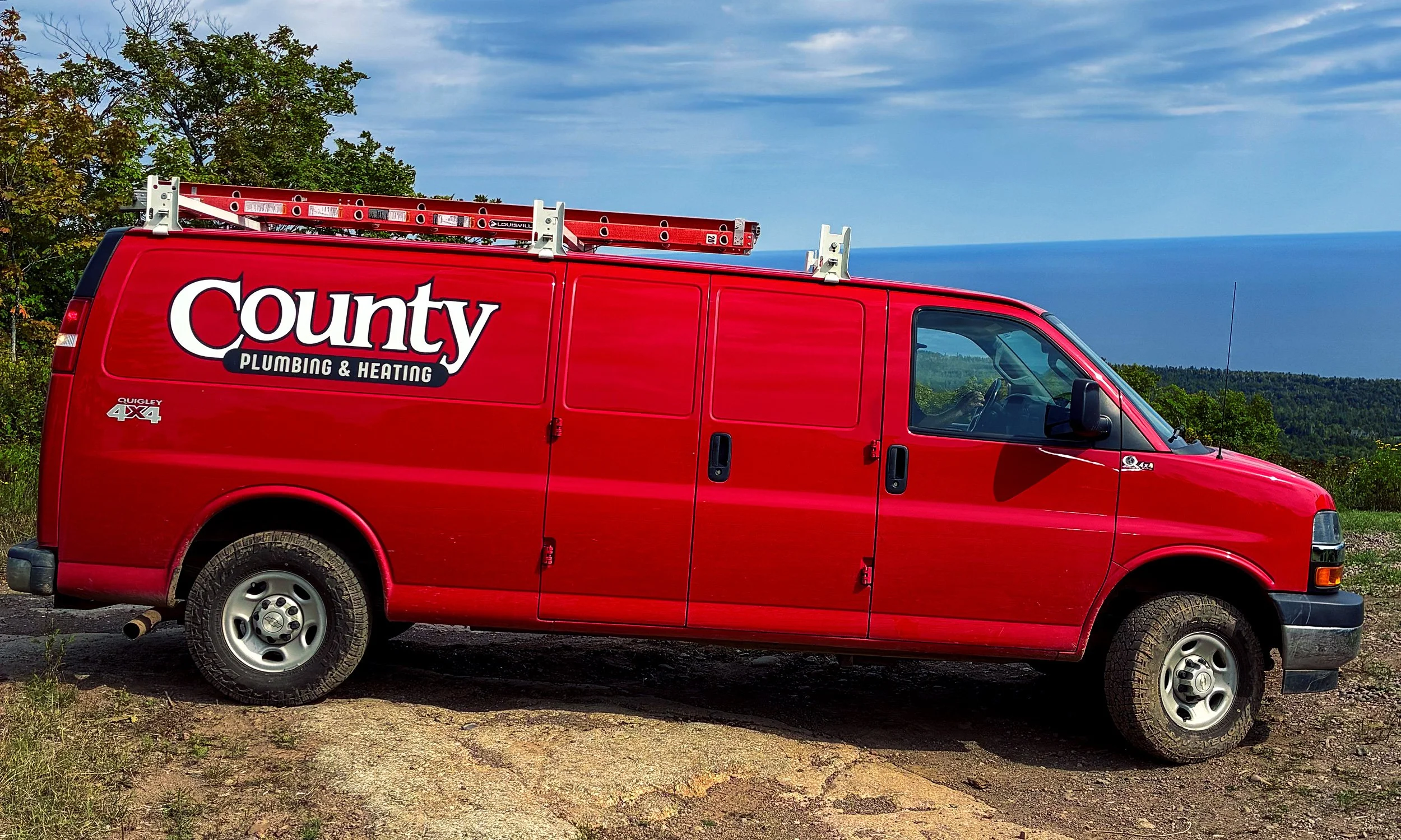 Red County Plumbing & Heating service van parked outdoors with a distant view of the Lake Superior in the background.