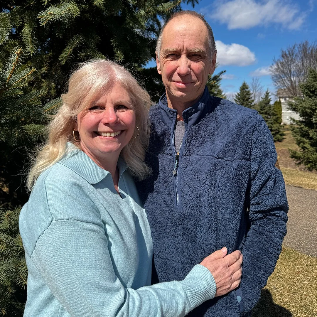 Tim & Tanya Goettl outdoors on a sunny day, standing in front of evergreen trees.