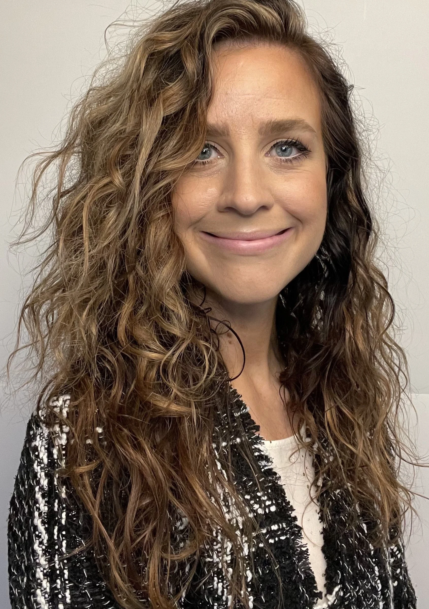 Close-up of a woman with wavy, shoulder-length brown hair, blue eyes, and a smile, wearing a black and white textured blazer. Laura Newmanfounder of Strong Culture