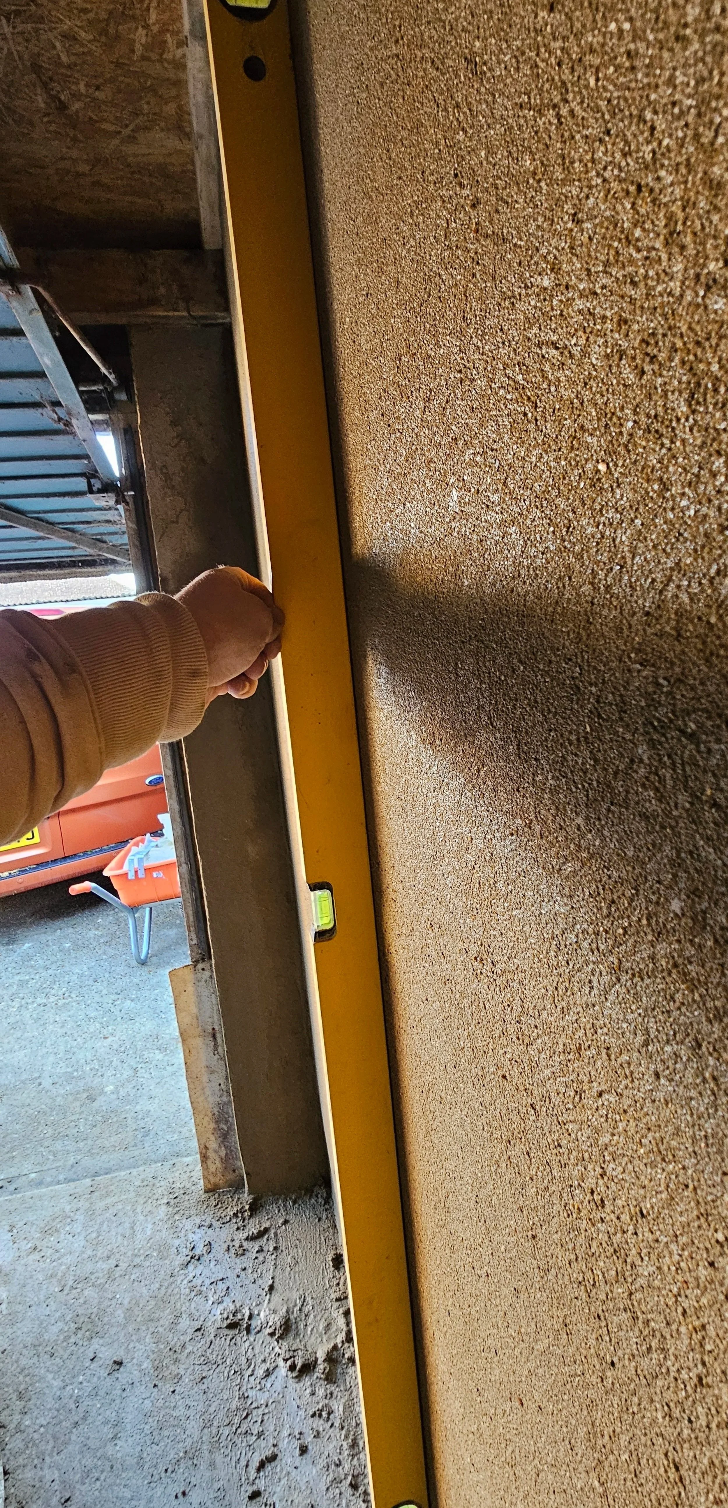 Person holding a yellow spirit level against a flat surface, indoors, with a concrete floor and construction materials visible.