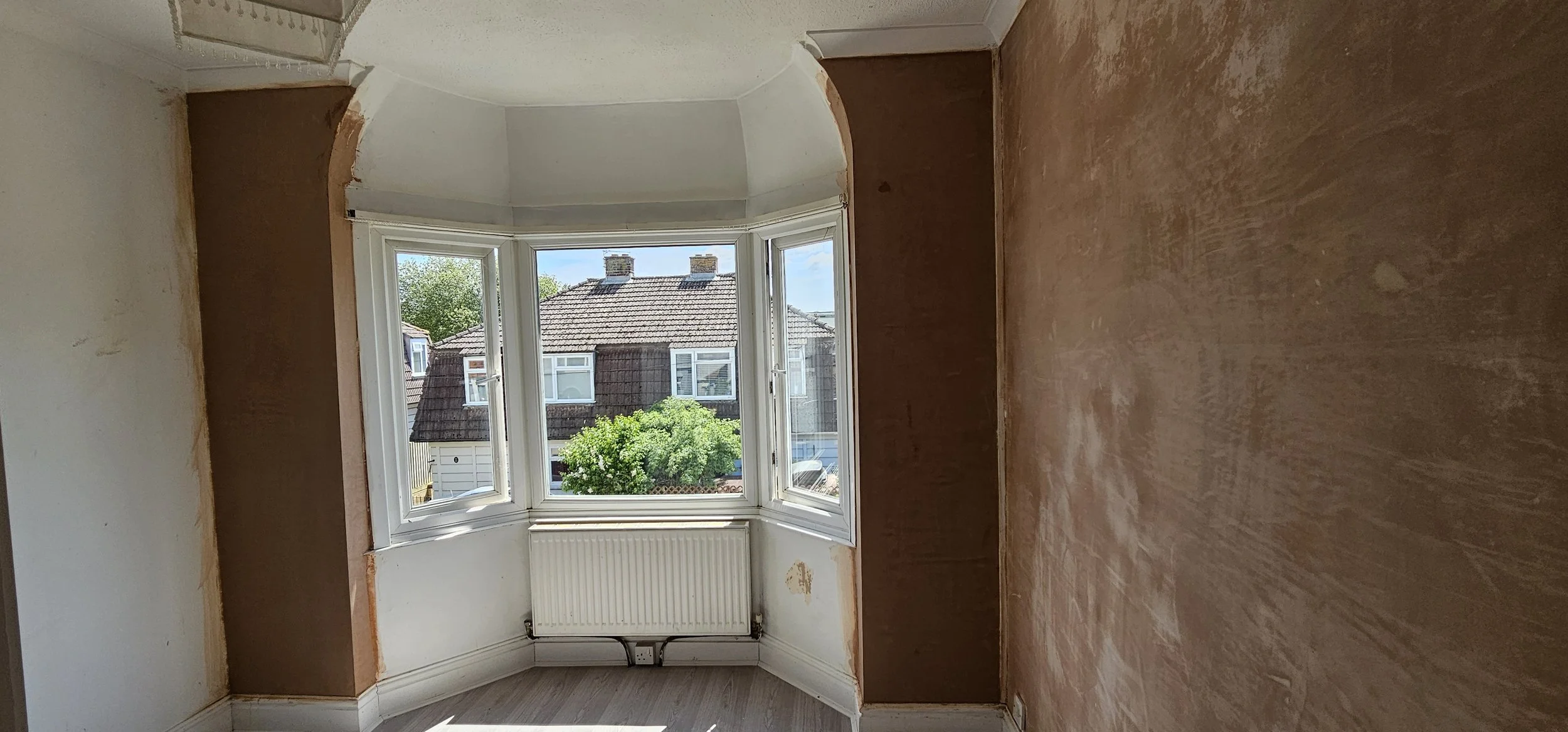 Room with large bay window overlooking houses and trees, partially painted walls, and a radiator below the window.