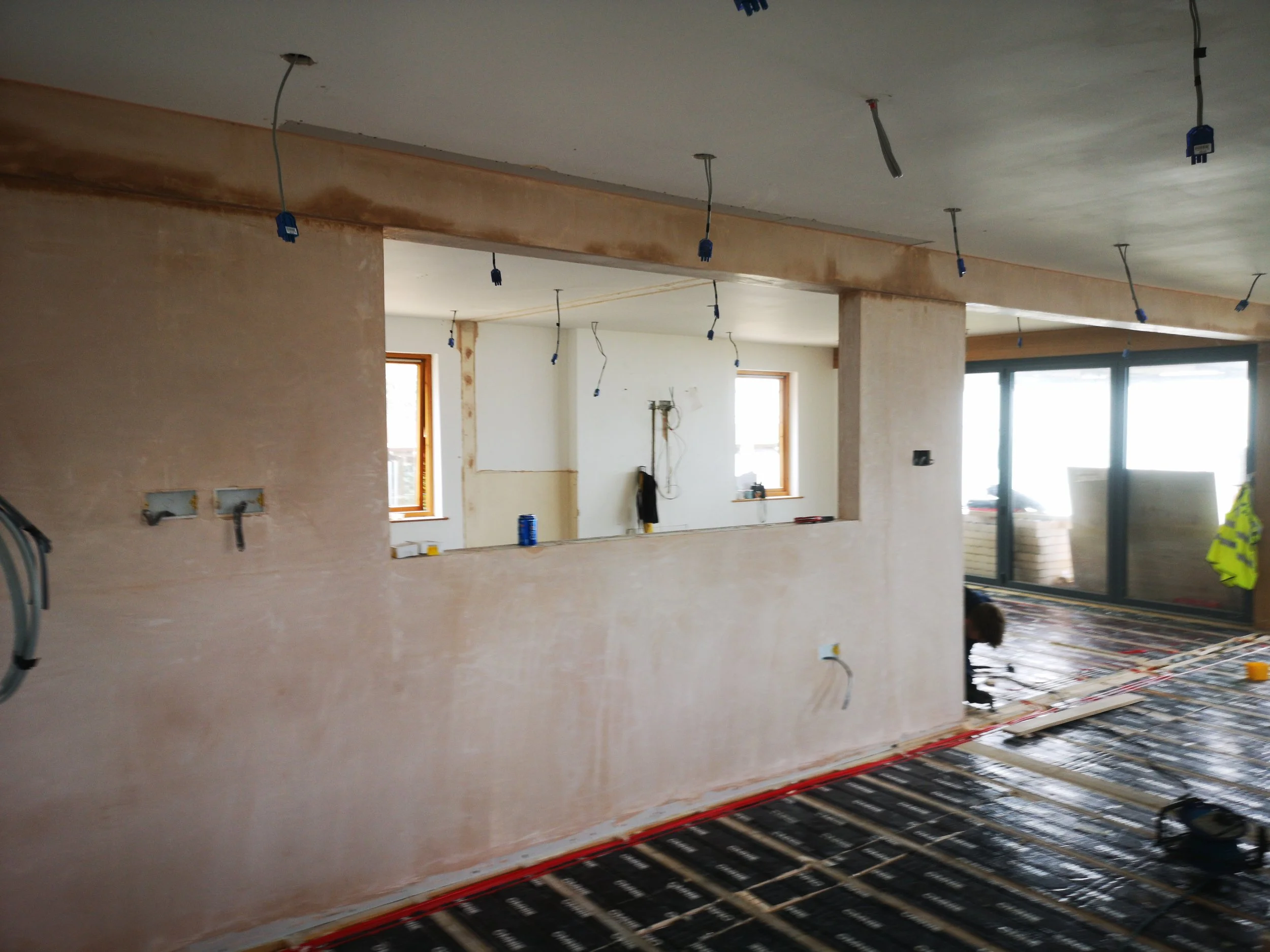Interior view of a house under construction with unfinished walls, exposed electrical wiring, large windows, and radiant floor heating installation.