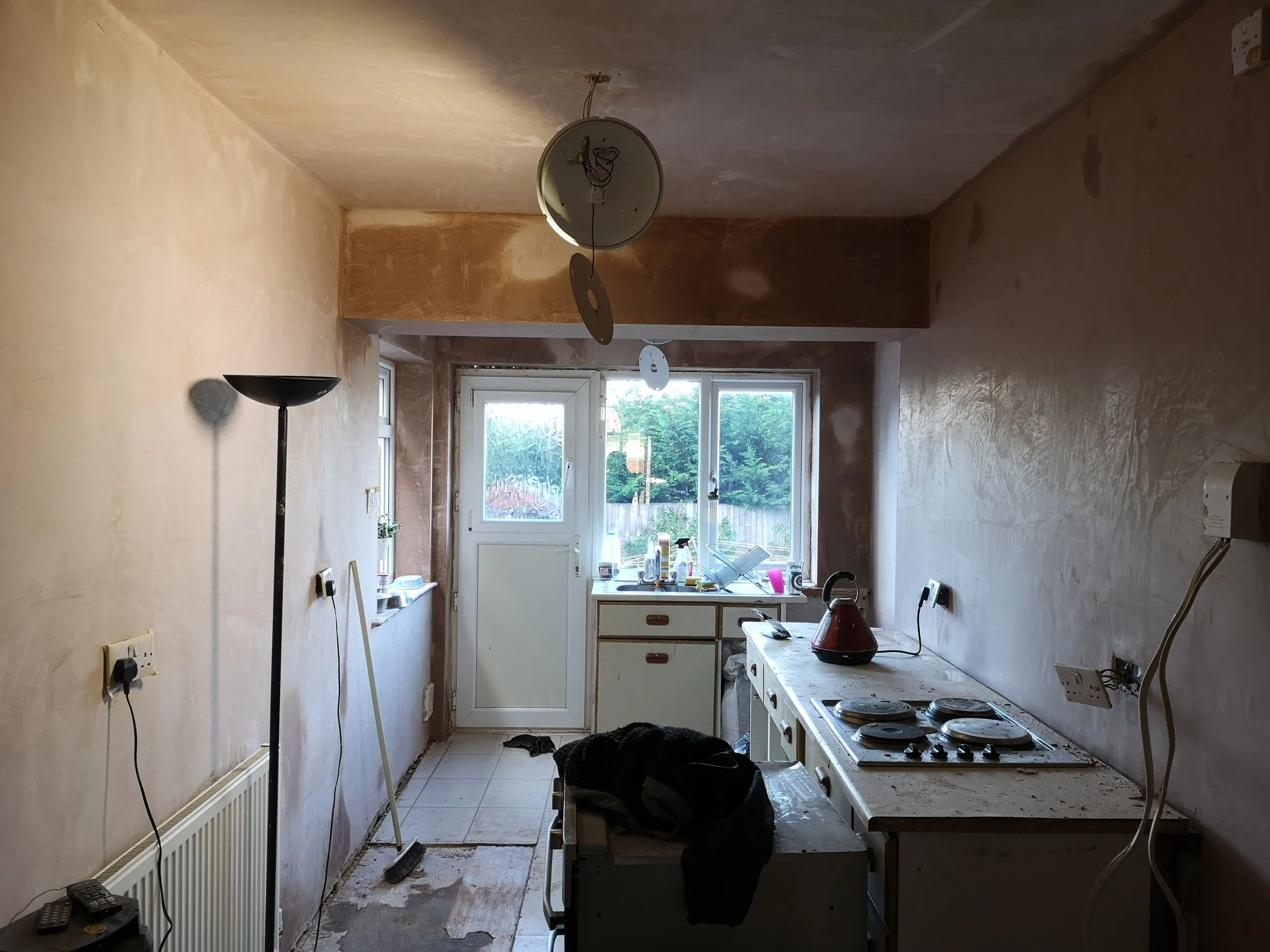 A kitchen undergoing renovation with unfinished walls, an old stovetop, a red kettle, a small sink, and a door leading outside. There is a tall floor lamp, debris on the floor, and construction tools.
