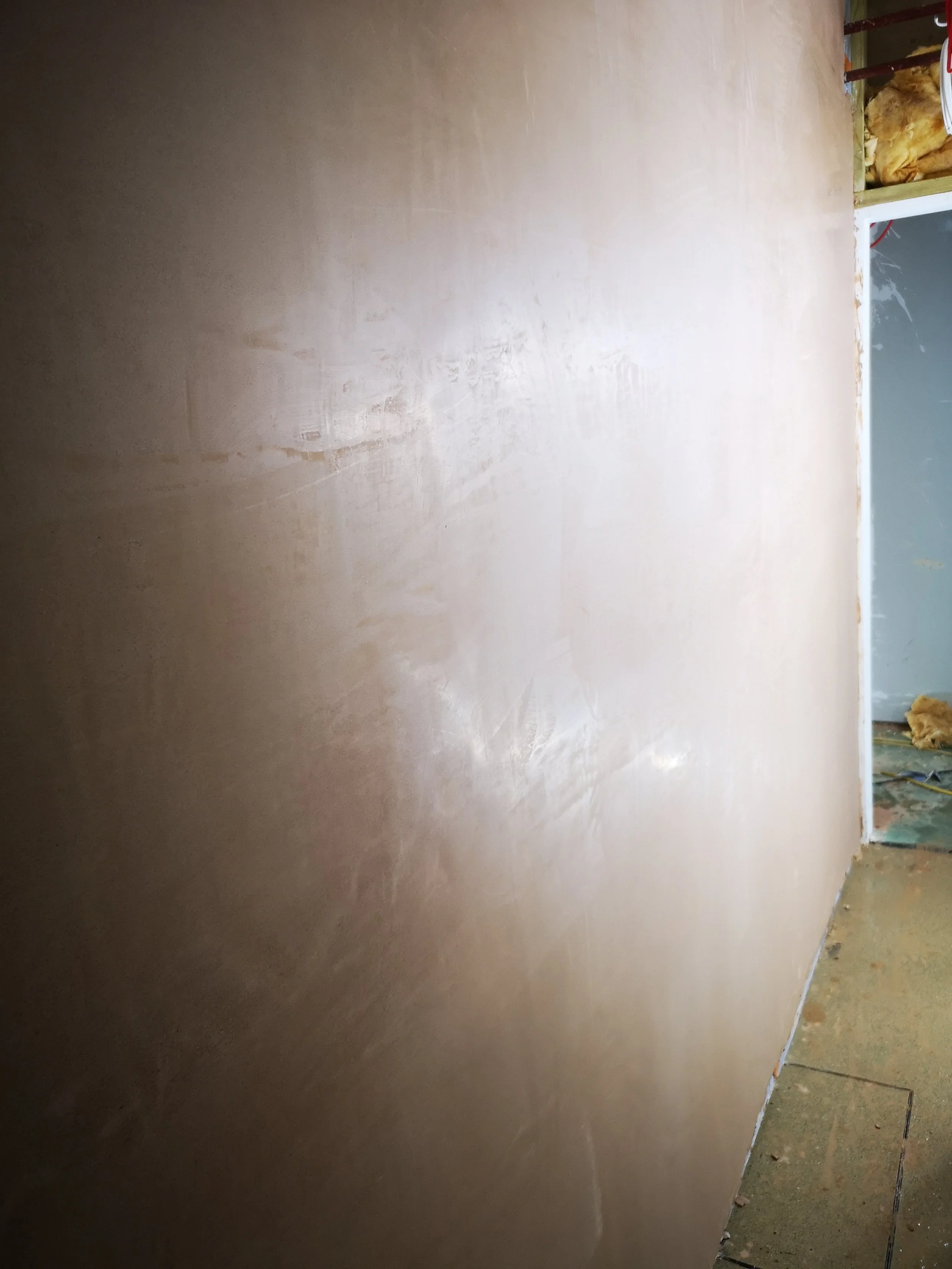 A freshly drywall wall inside a building under construction, with visible seams and joint compound, and construction materials visible at the corner.