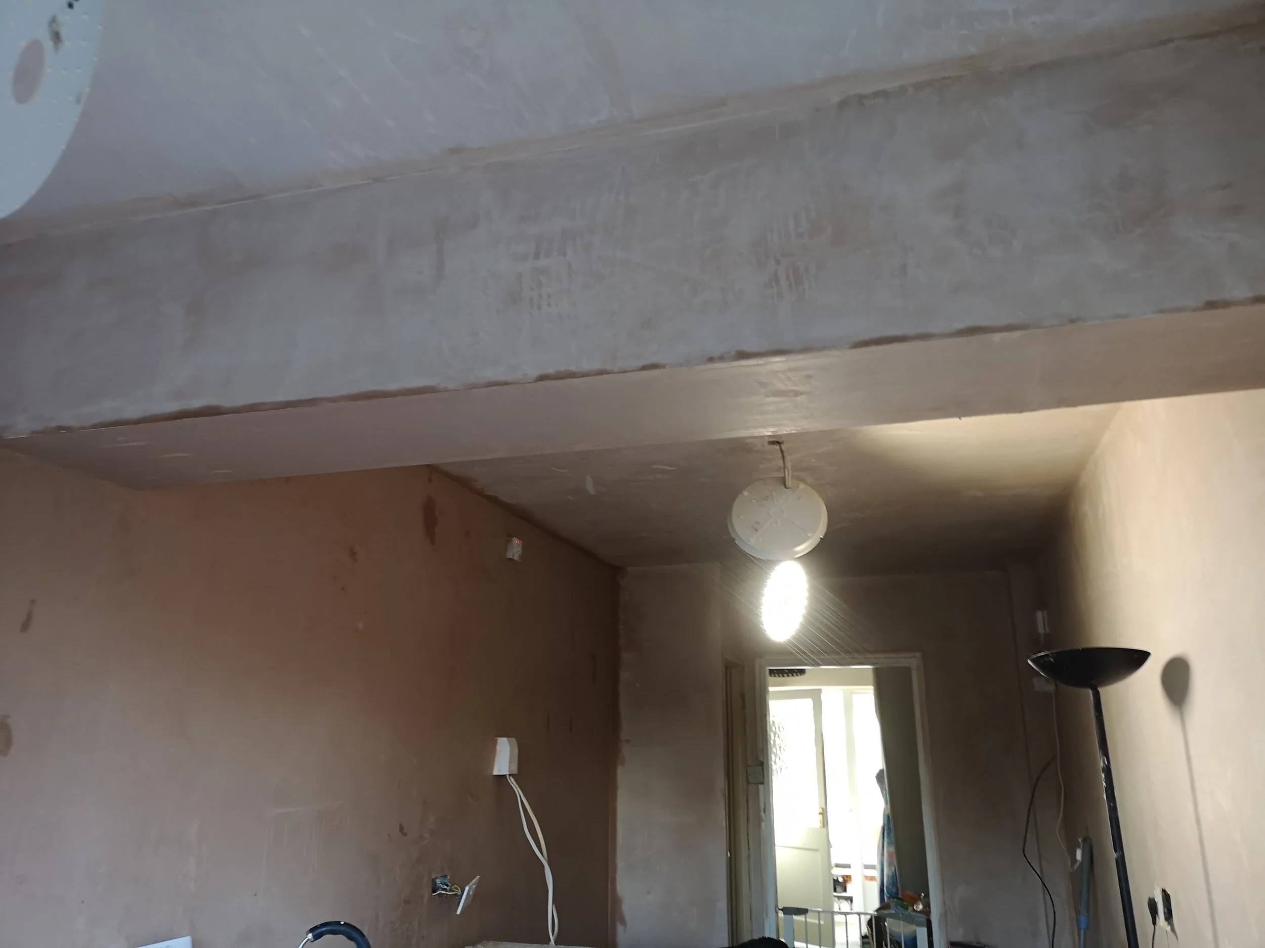 Interior of a room under construction or renovation with unfinished walls, a ceiling light fixture, and a standing lamp. There are visible wires and electrical outlets on the walls, and a doorway leading to another room.