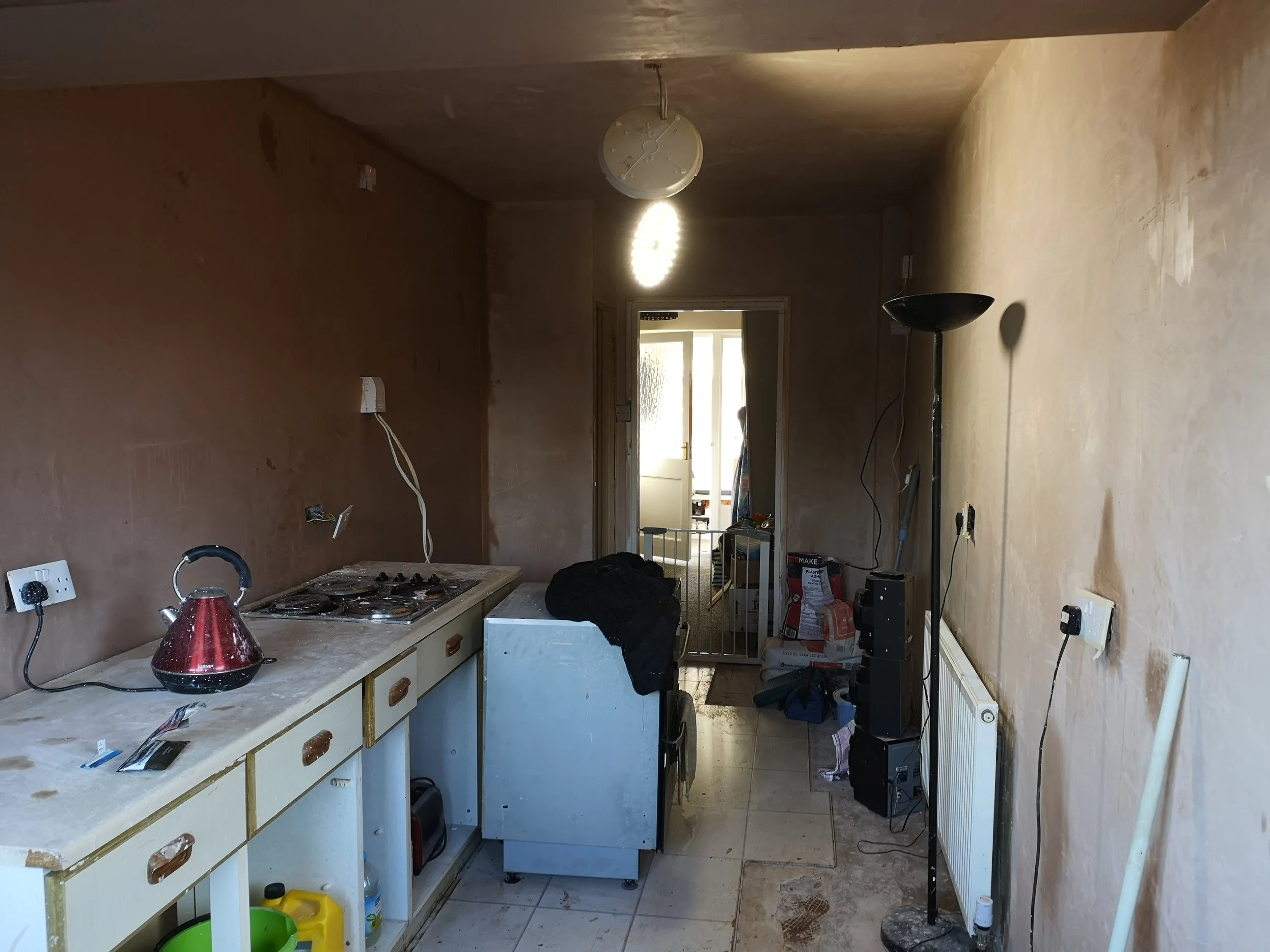 A cluttered kitchen with an unclean countertop, a red kettle, and outdated cabinets. There is a stove, a washing machine, a standing lamp, and some household items scattered around, with unfinished walls and a doorway leading to another room.