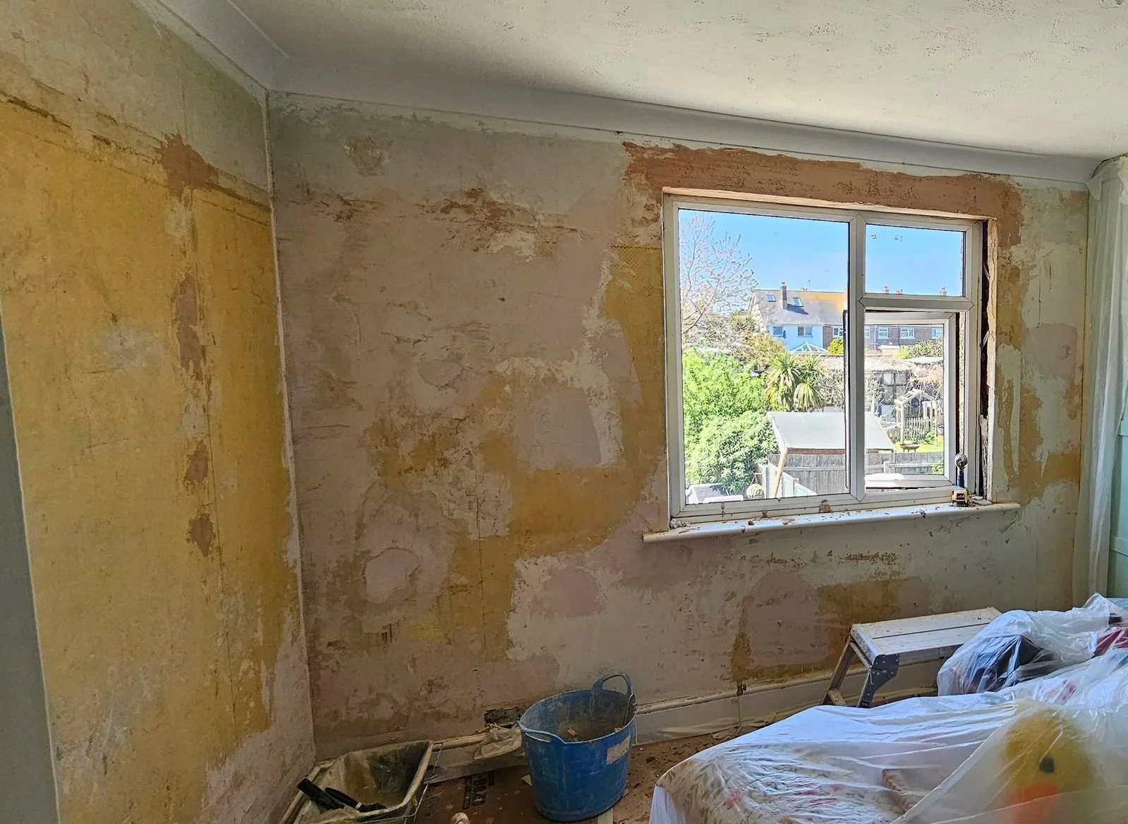 Room undergoing renovation with distressed yellow and beige walls, a window showing a backyard with trees and houses, and renovation tools and materials on the floor.