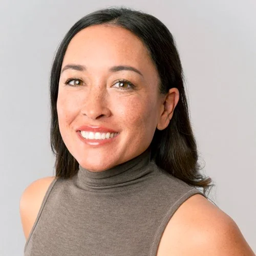 A woman with shoulder-length dark hair, freckles, and light skin color, smiling widely with teeth showing, wearing a light pink button-up shirt and a dangling earring.