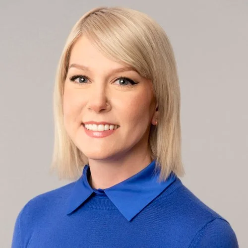 A woman with short blonde hair, wearing a blue collared sweater, smiling at the camera against a light grey background.