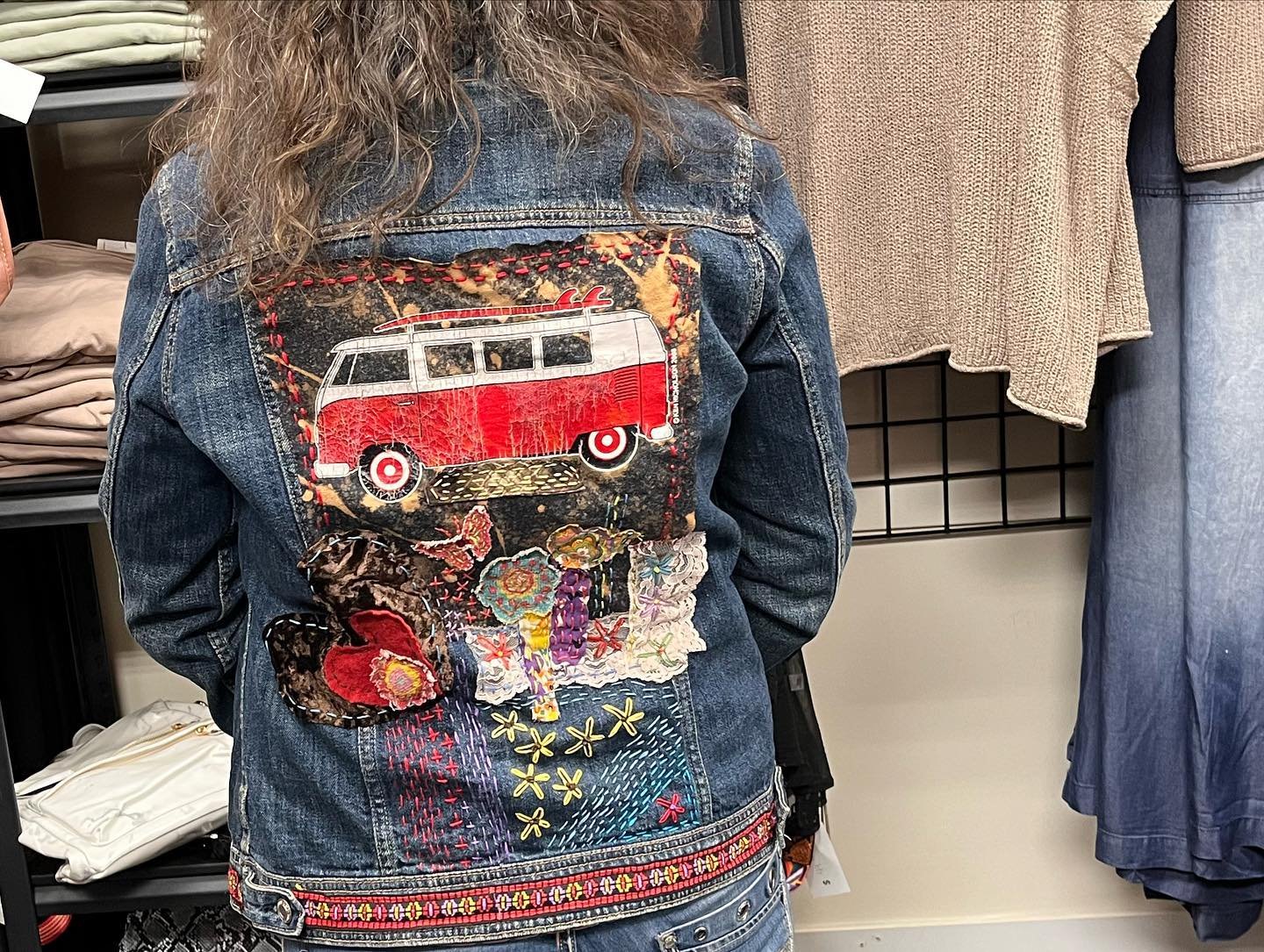 Back of a woman wearing a denim jacket with a patchwork of embroidered and fabric patches featuring a red vintage van, flowers, and colorful patterns