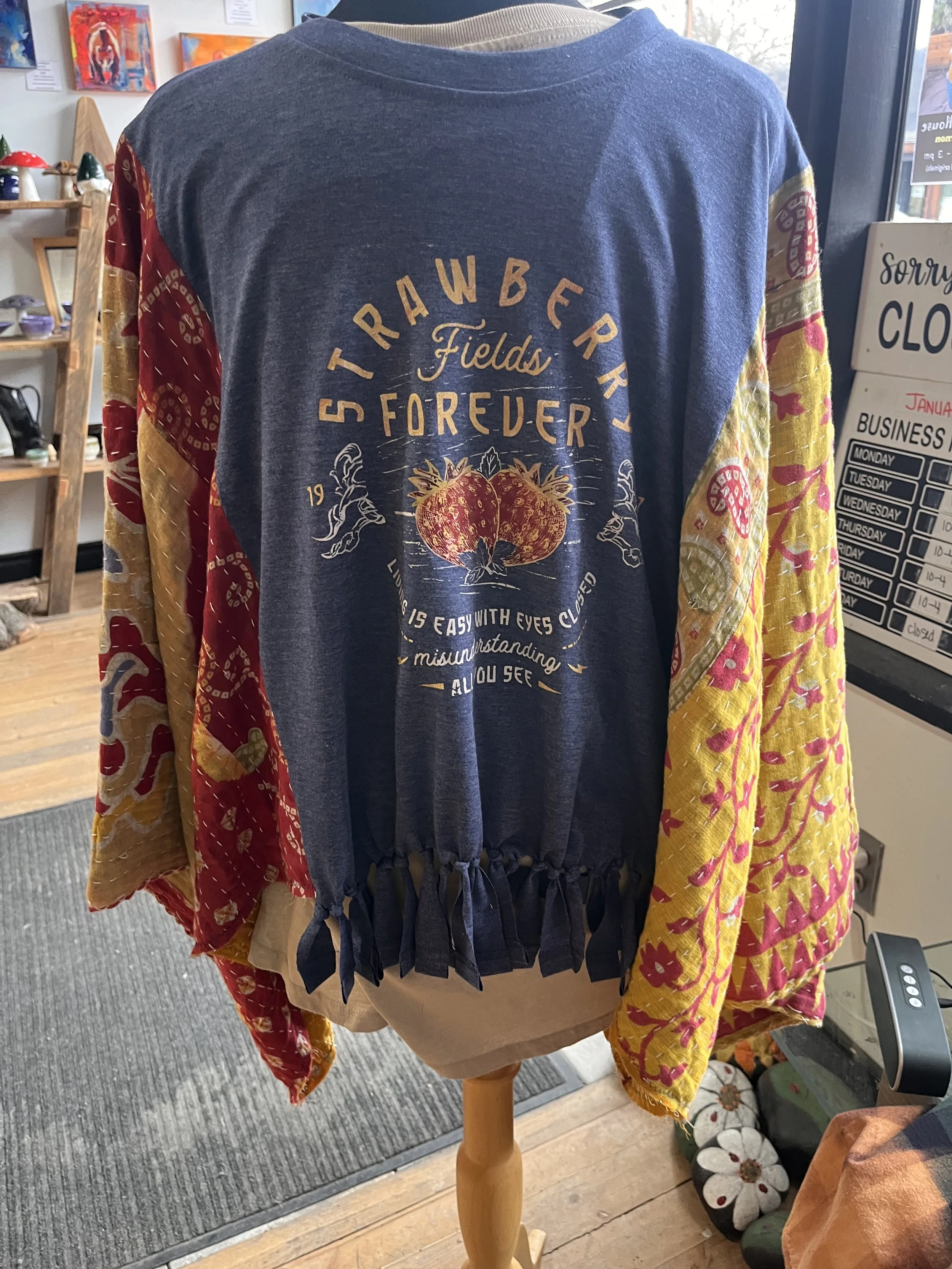 A shirt displayed on a mannequin with colorful sleeves. The back of the shirt has a graphic design featuring strawberries and the text 'Strawberry Fields Forever' with additional decorative text and symbols.