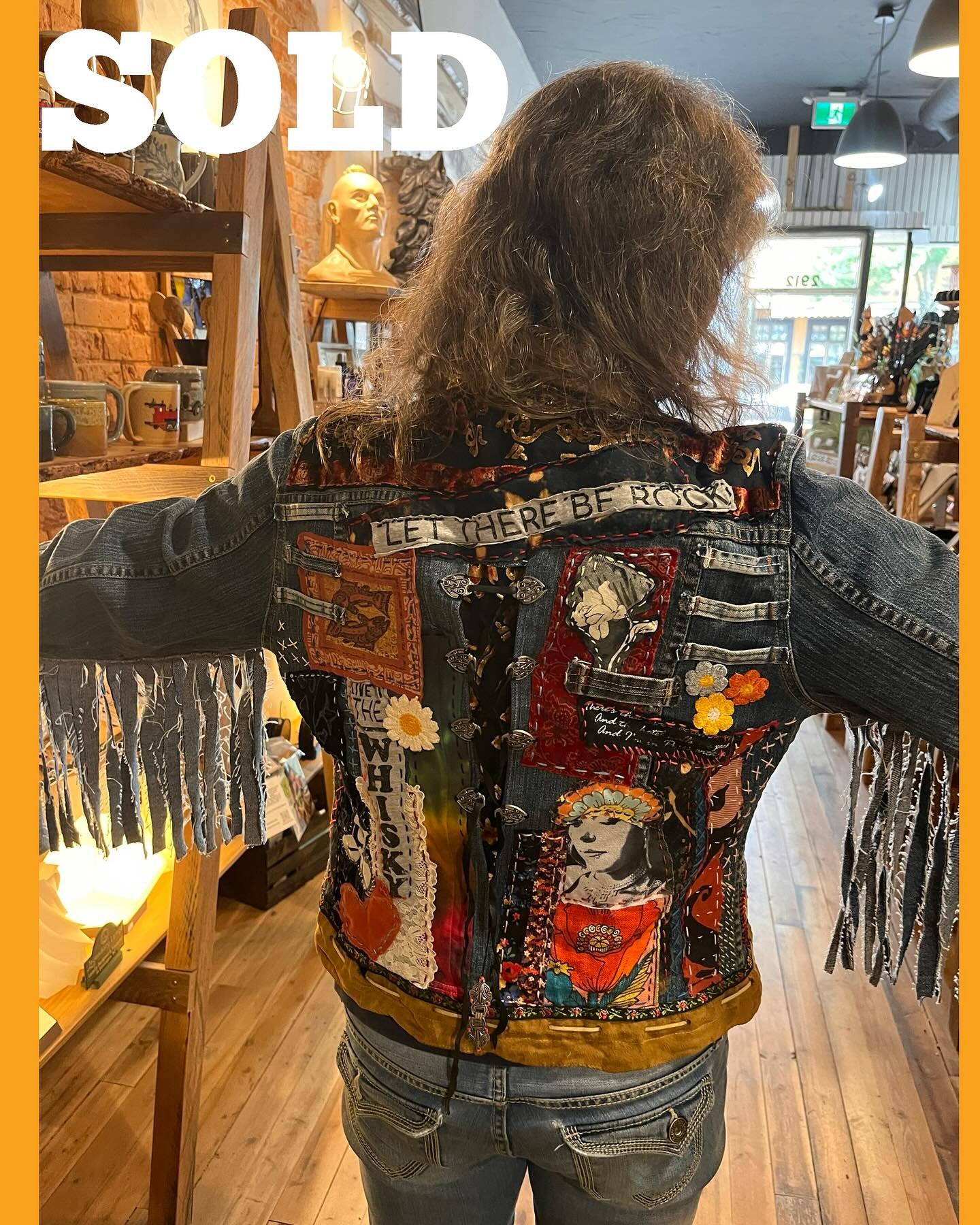 Person wearing a colorful, patchwork denim jacket with fringe on the sleeves inside a store. The jacket has various patches, embroidery, and text such as "LET THERE BE ROCK". The store has wooden shelves, decorative busts, and various items on displa