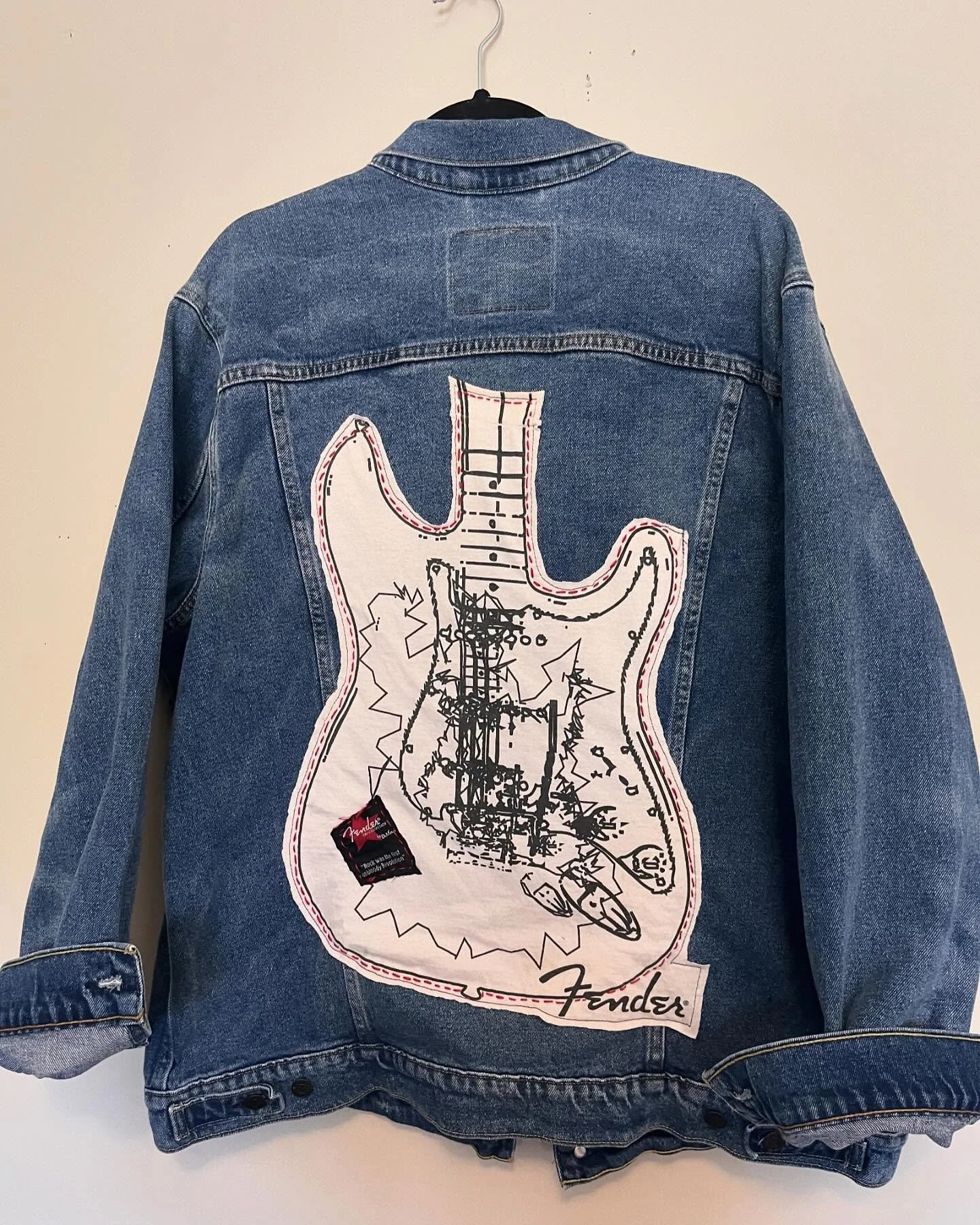A blue denim jacket with a large embroidered patch of a guitar on the back, featuring sketch-style art, a small black tag, and the Fender logo at the bottom.