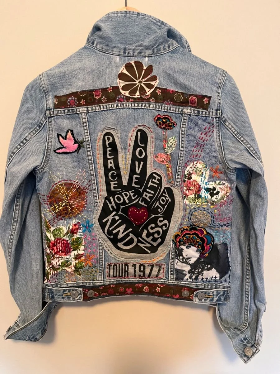 Back of a light blue denim jacket decorated with embroidered patches, including a large peace sign with words, flowers, butterflies, and a portrait of a woman with colorful hair, and a patch with "Tour 1977".