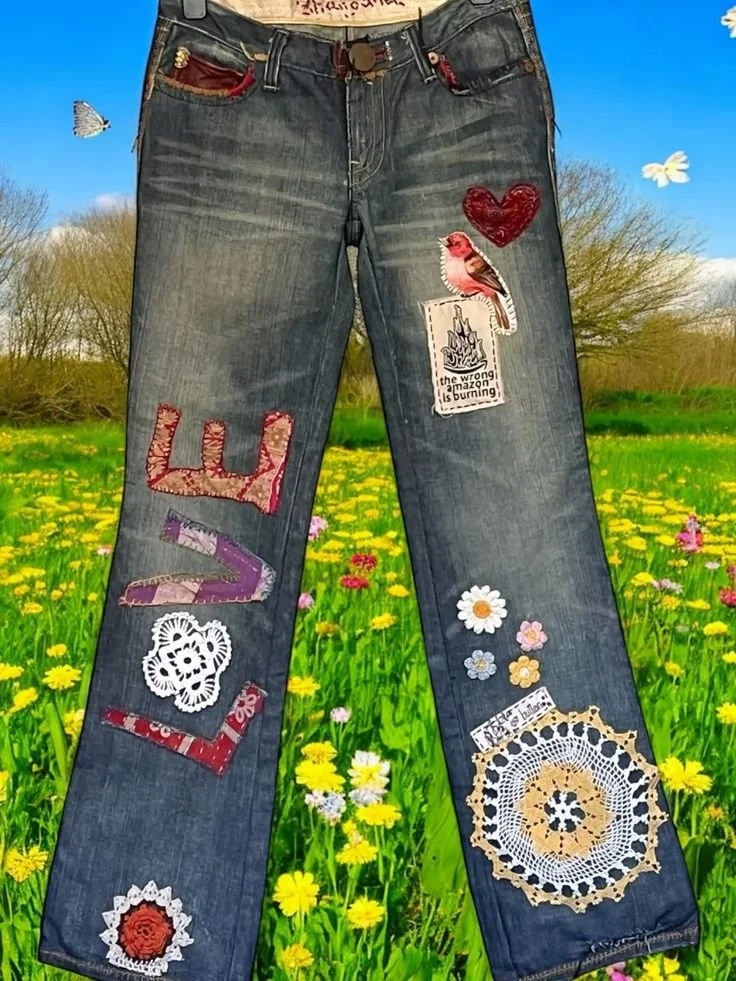 Pair of denim jeans decorated with various patches including a red heart, a pink bird, a white lace face, and colorful floral patterns, hanging against a scenic background of a yellow flower field and blue sky.