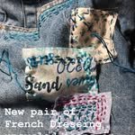 Close-up of fabric with beach sand, a tag, and French dress-themed embroidery or print.