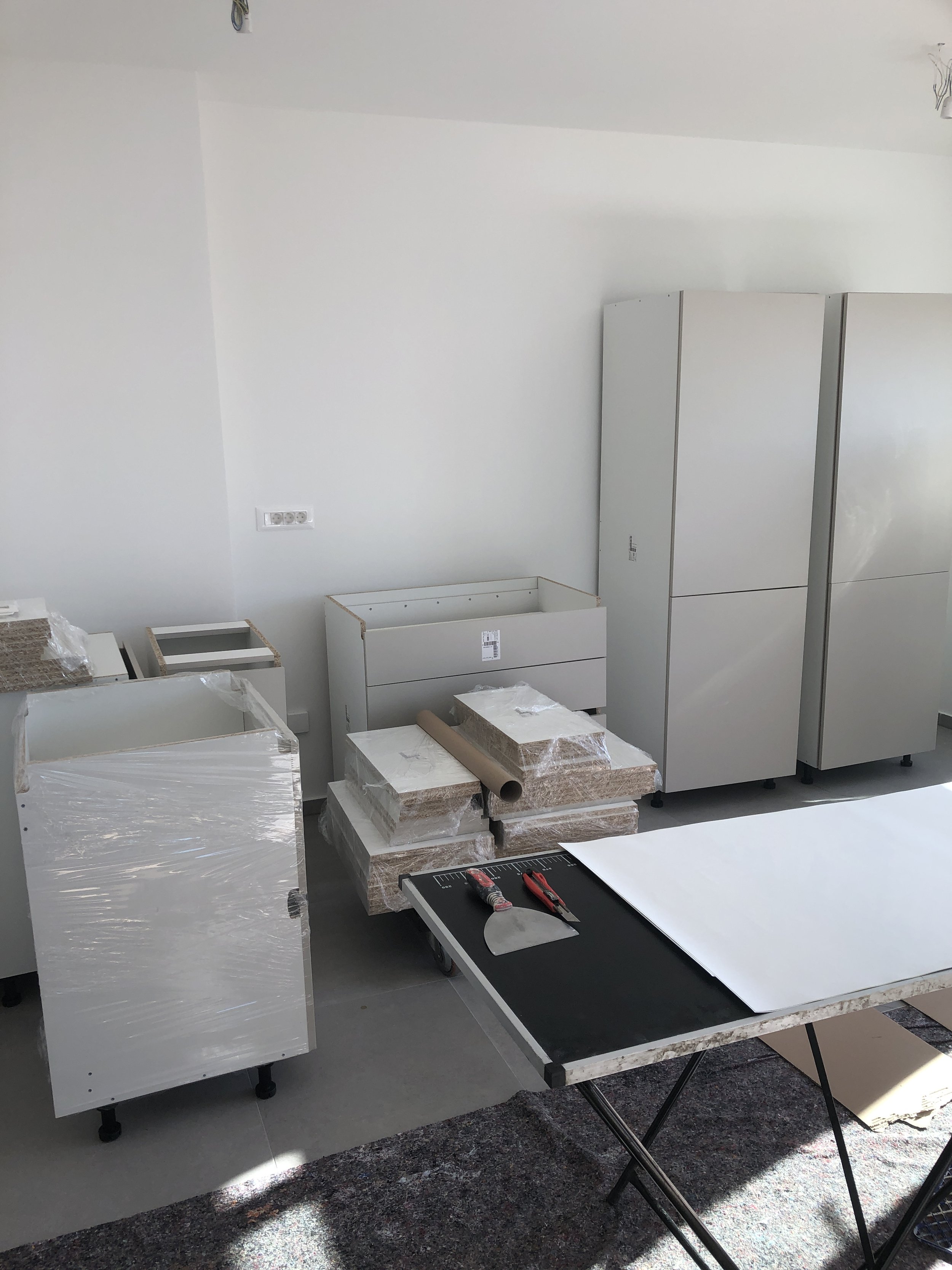 Soba u izgradnji sa sastavljenim bijelim namještajem, građevinskim alatima i materijalima za pakiranje, uključujući sklopivi stol, omotane ormariće i radni prostor.