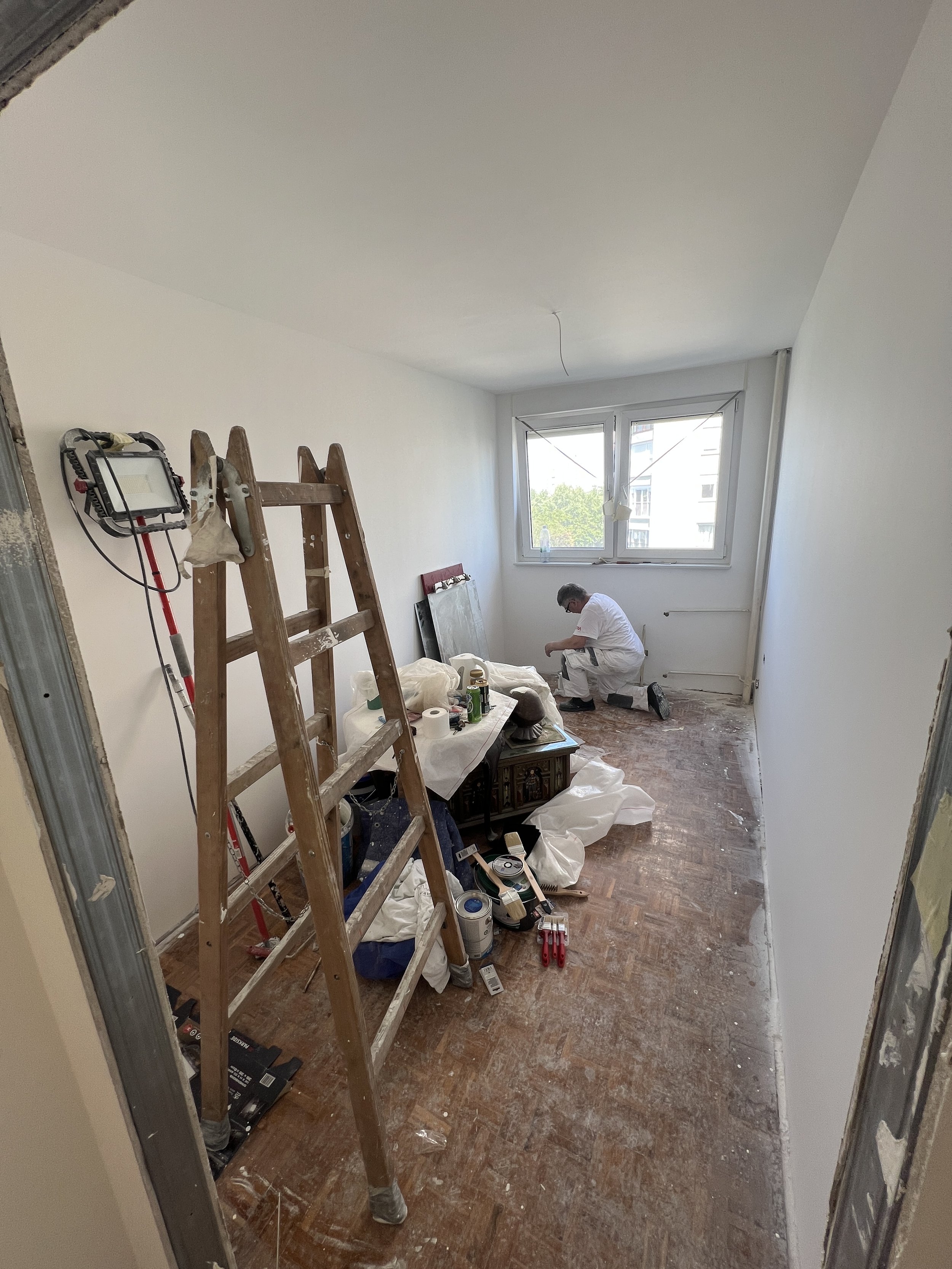 Osoba koja radi na projektu obnove ili bojanja u maloj sobi s bijelim zidovima i prozorom, okružena alatima, limenkama boje i priborom.