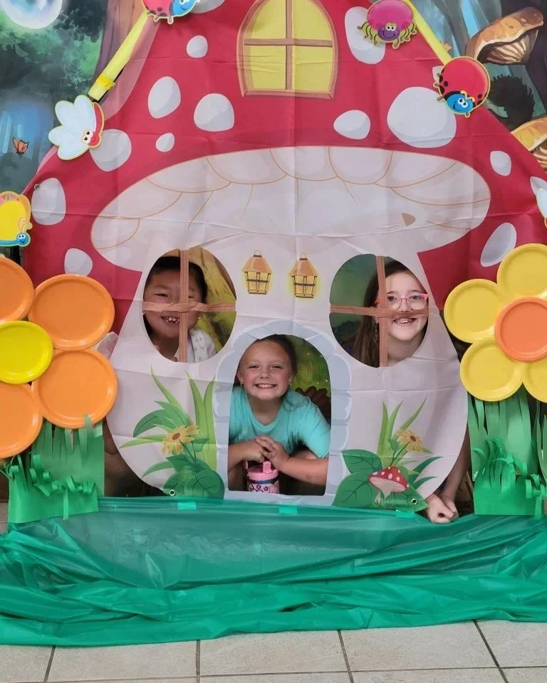 Three children peeking through cutouts in a colorful fairy tale mushroom house prop with flowers and bugs decoration.