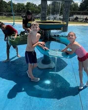 Children playing at a water park with a splash pad feature, children at a fountain, and a water play area in the background.