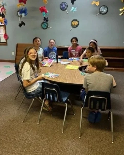 Children sitting around a table in a classroom or activity room, with colorful paper fans on the wall behind them.