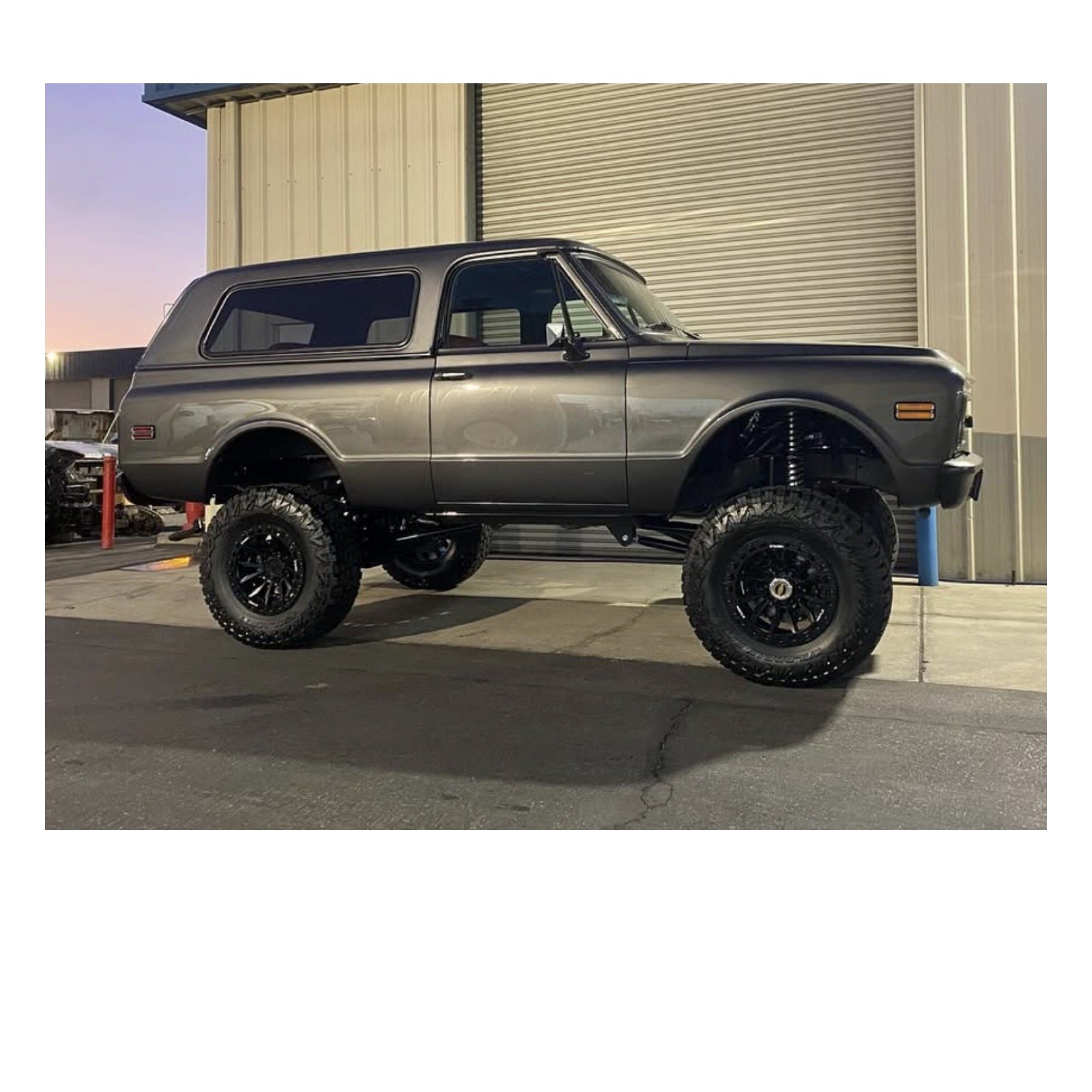 Gray lifted SUV with large off-road tires parked outside near a metal building at sunset.