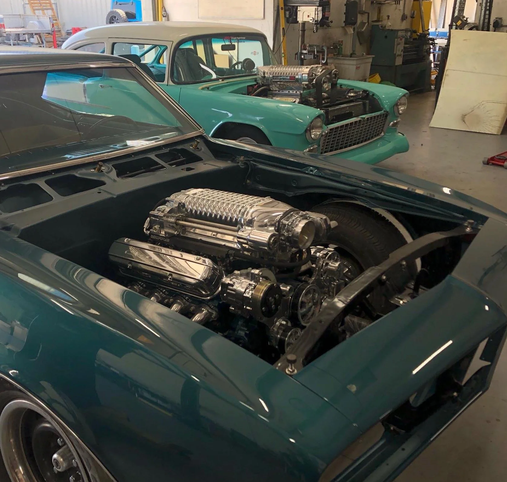 Two classic cars in a garage, one with a visible high-performance engine, and another with a small engine, both undergoing restoration or maintenance.