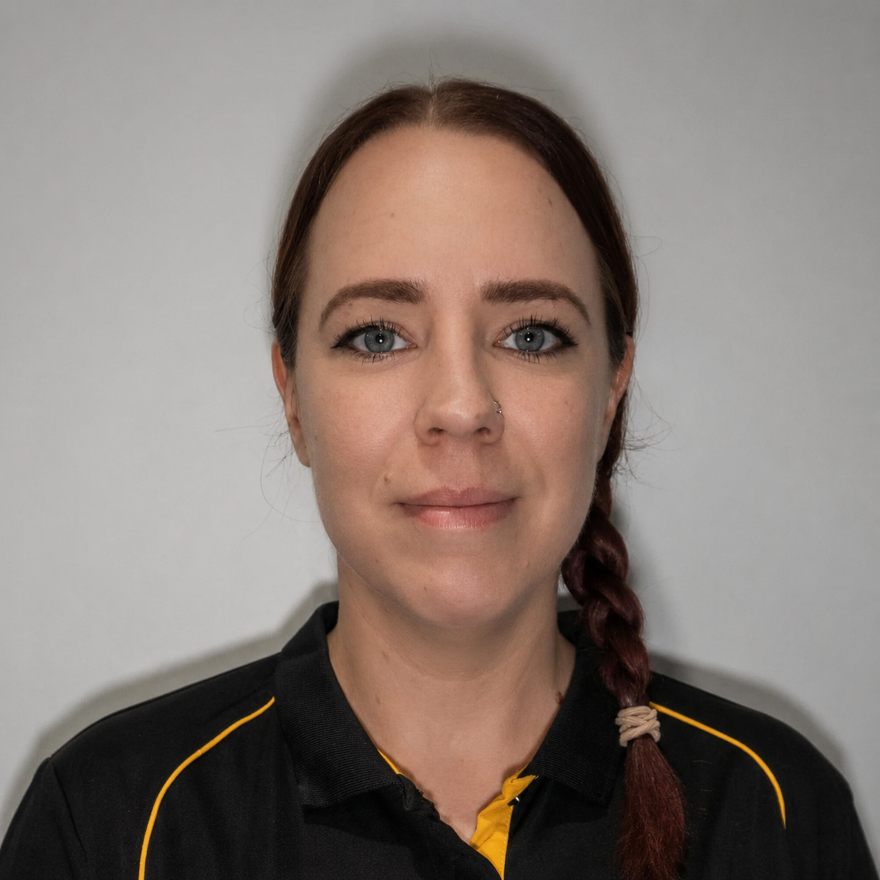 A woman with blue eyes, red hair in a braid, wearing a black shirt with yellow accents, standing against a plain gray background.