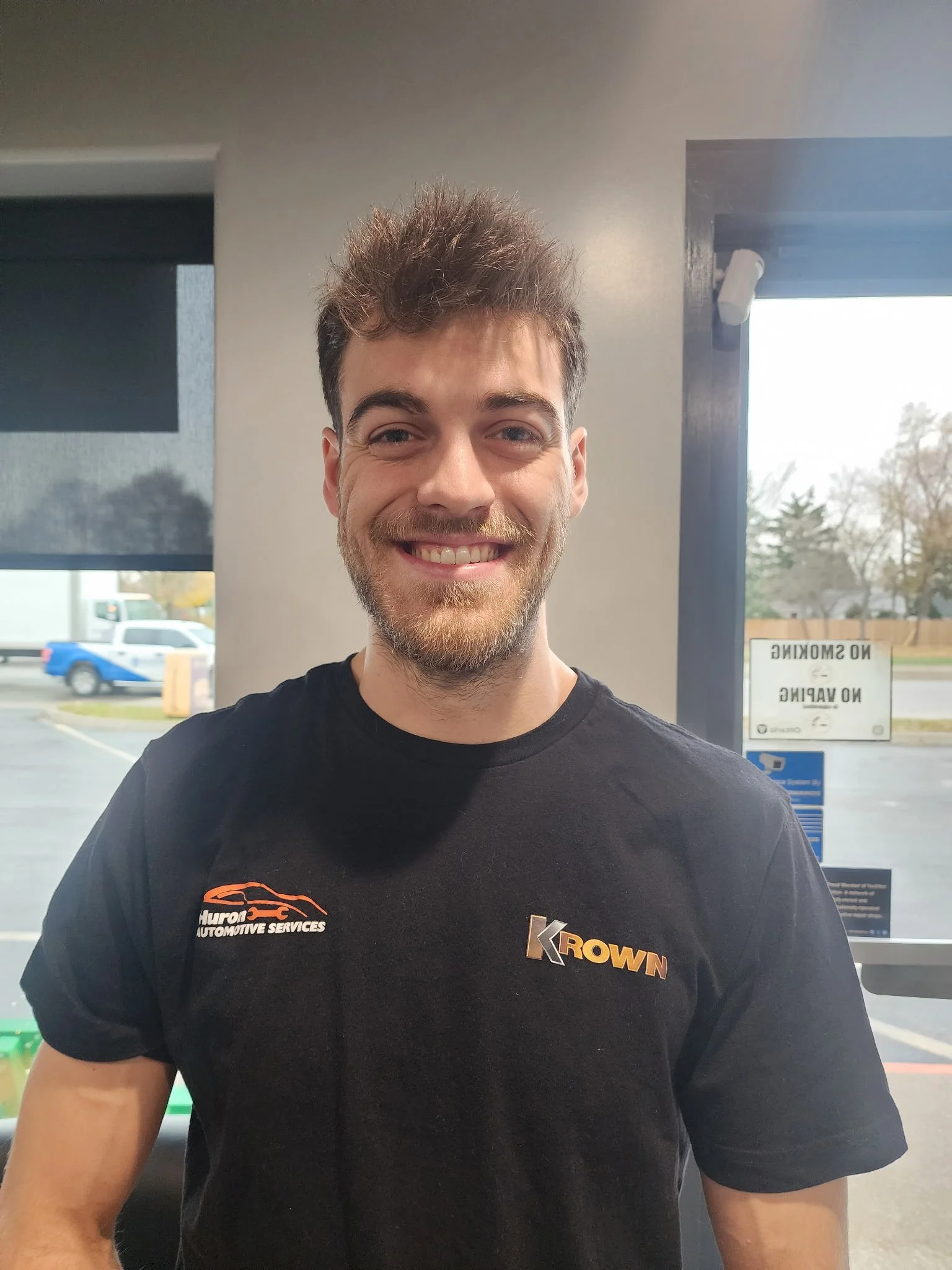 A young man with a beard and short, tousled brown hair smiles at the camera. He is wearing a black T-shirt with orange and white text and graphics for 'Hurón Automotive Services' and 'Krown.' The background shows a window with a parking lot outside, where a blue and white vehicle is visible, and trees and cloudy sky beyond.