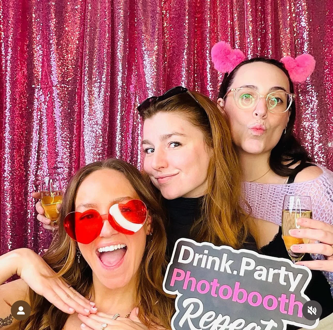 Three women at a celebration with a pink, sparkly backdrop, wearing fun accessories, holding drinks, and smiling. One woman is wearing red heart-shaped glasses and has a big open-mouthed smile, another has sunglasses on her head and is making a playf