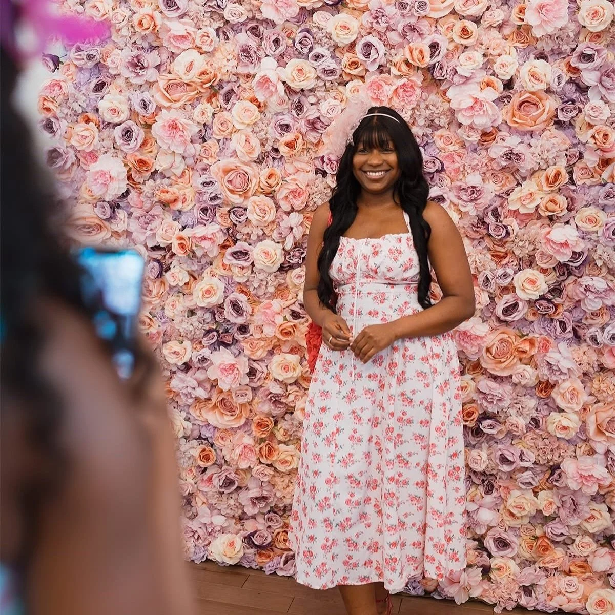Woman in a floral dress standing in front of a pink and purple flower wall, smiling.