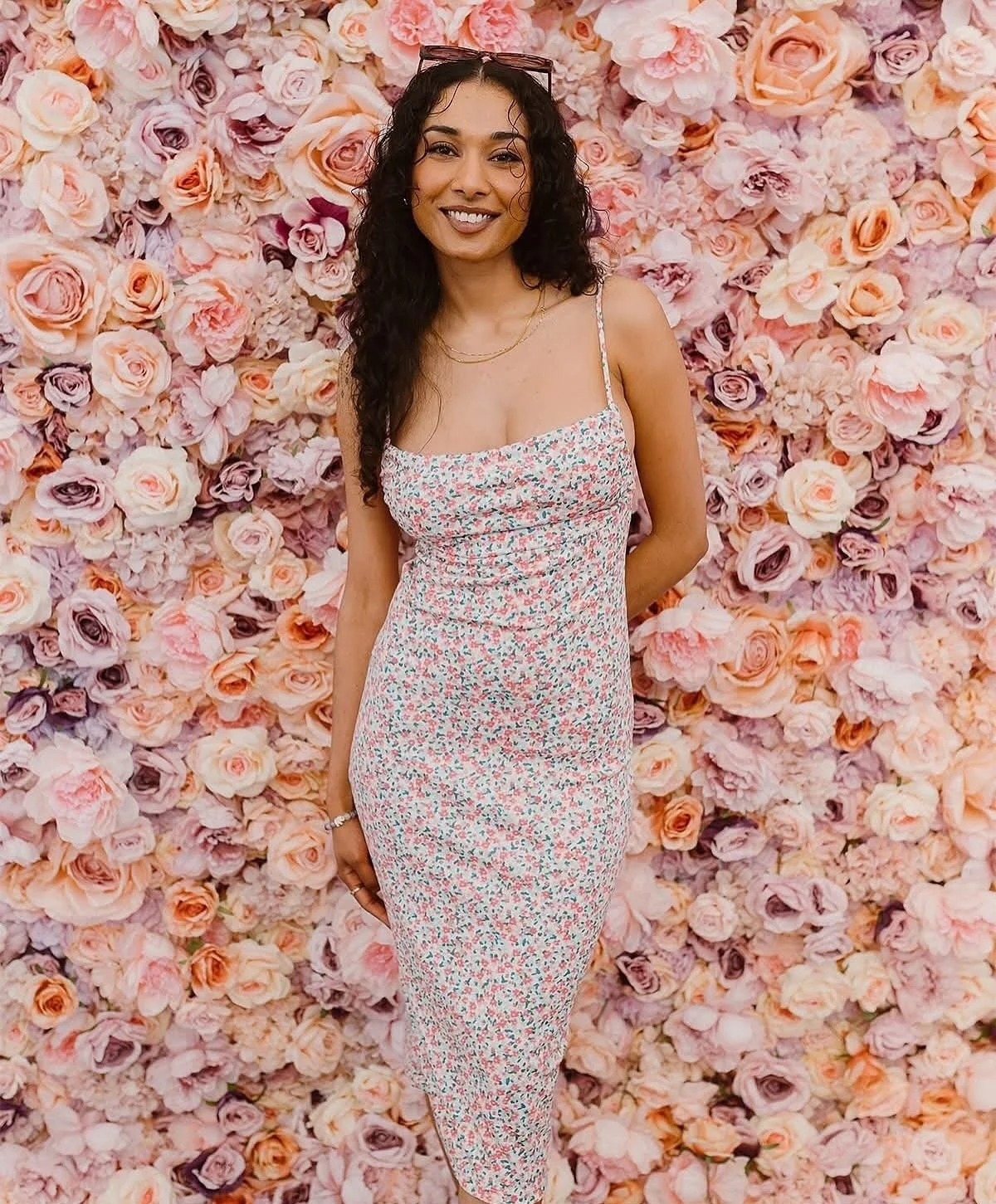 A woman with long, black curly hair and sunglasses on her head stands in front of a wall adorned with pink and peach roses. She is smiling and wearing a sleeveless, floral-patterned dress.