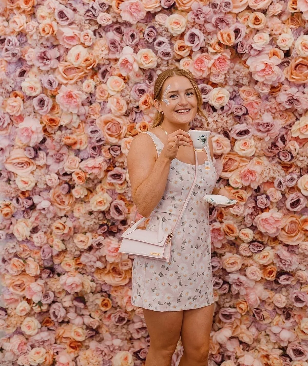 Woman in a floral dress standing in front of a wall covered in pink and purple roses, holding a teacup and saucer, smiling.