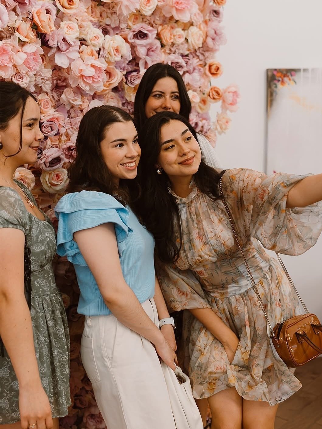 Group of five women taking a selfie in front of a wall decorated with pink and peach flowers at a social event.