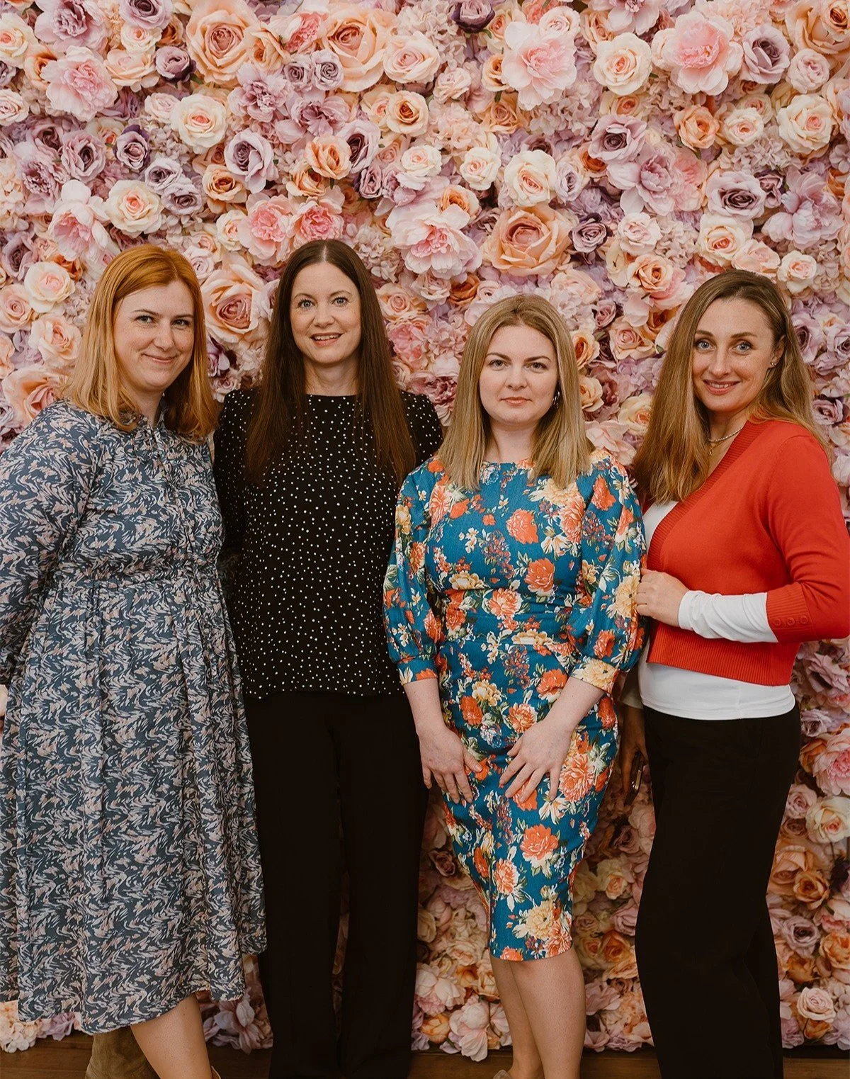 Four women standing in front of a floral pink and purple flower wall, wearing colorful dresses and smiling.