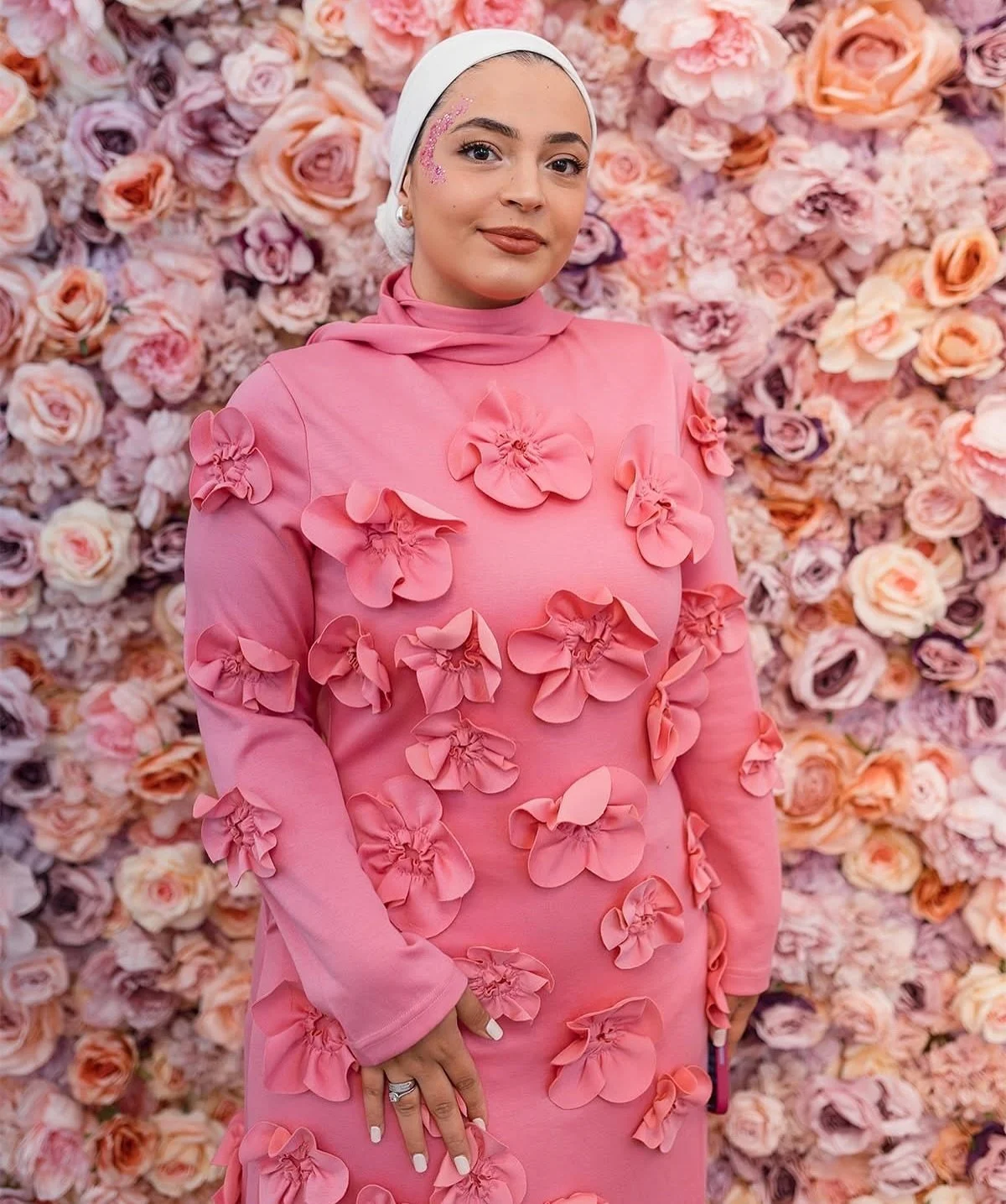A woman wearing a pink dress with 3D floral embellishments stands in front of a backdrop made of pink and peach roses, wearing a white headscarf and makeup with pink floral face decorations.