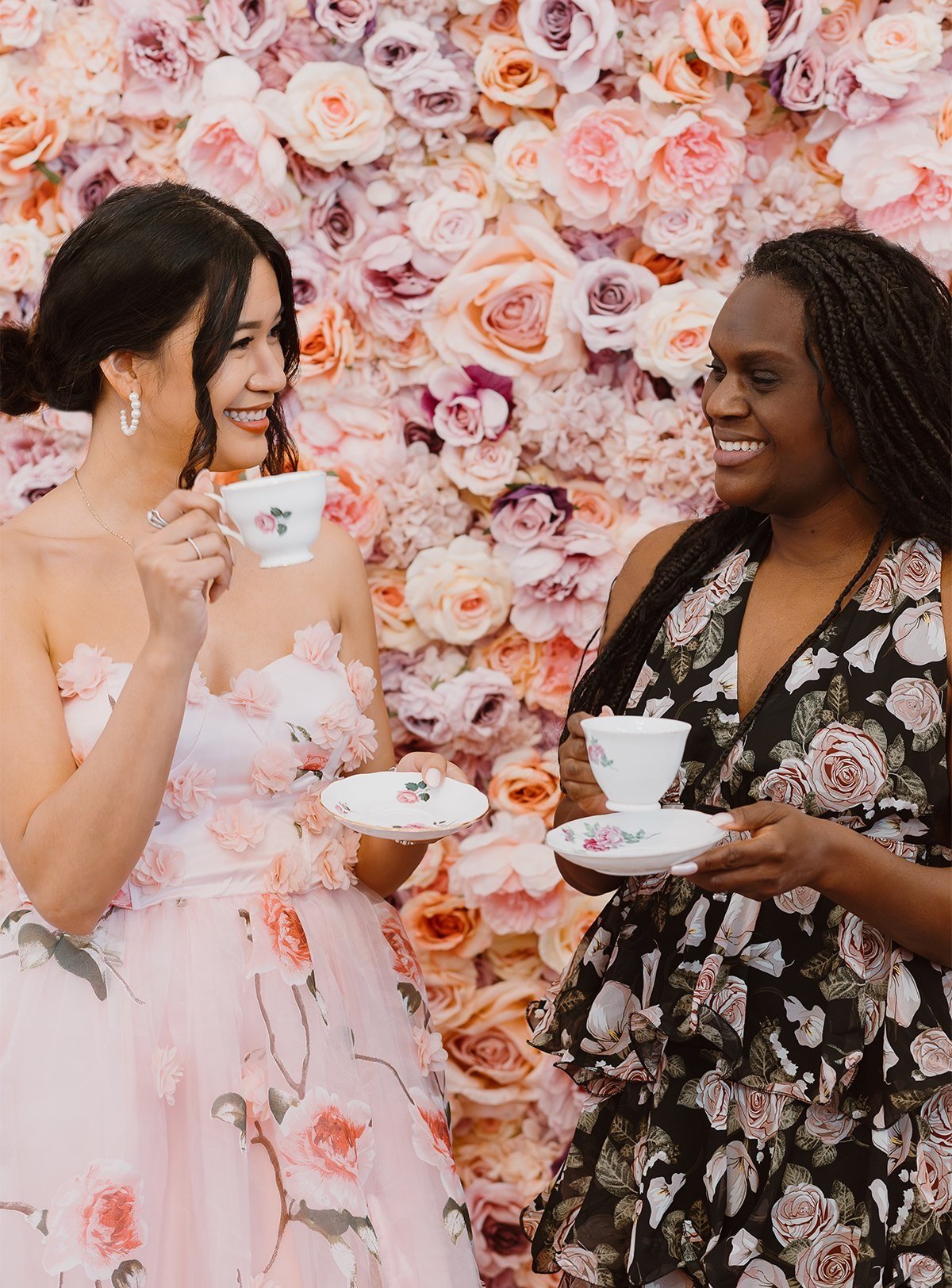 Two women in floral dresses holding teacups and saucers, smiling and chatting in front of a pink and peach floral background.